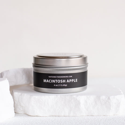 A 4 oz metal tin candle labeled 'MACINTOSH APPLE' by Nature's Craft and More, sitting on a white stone pedestal against a clean white background.