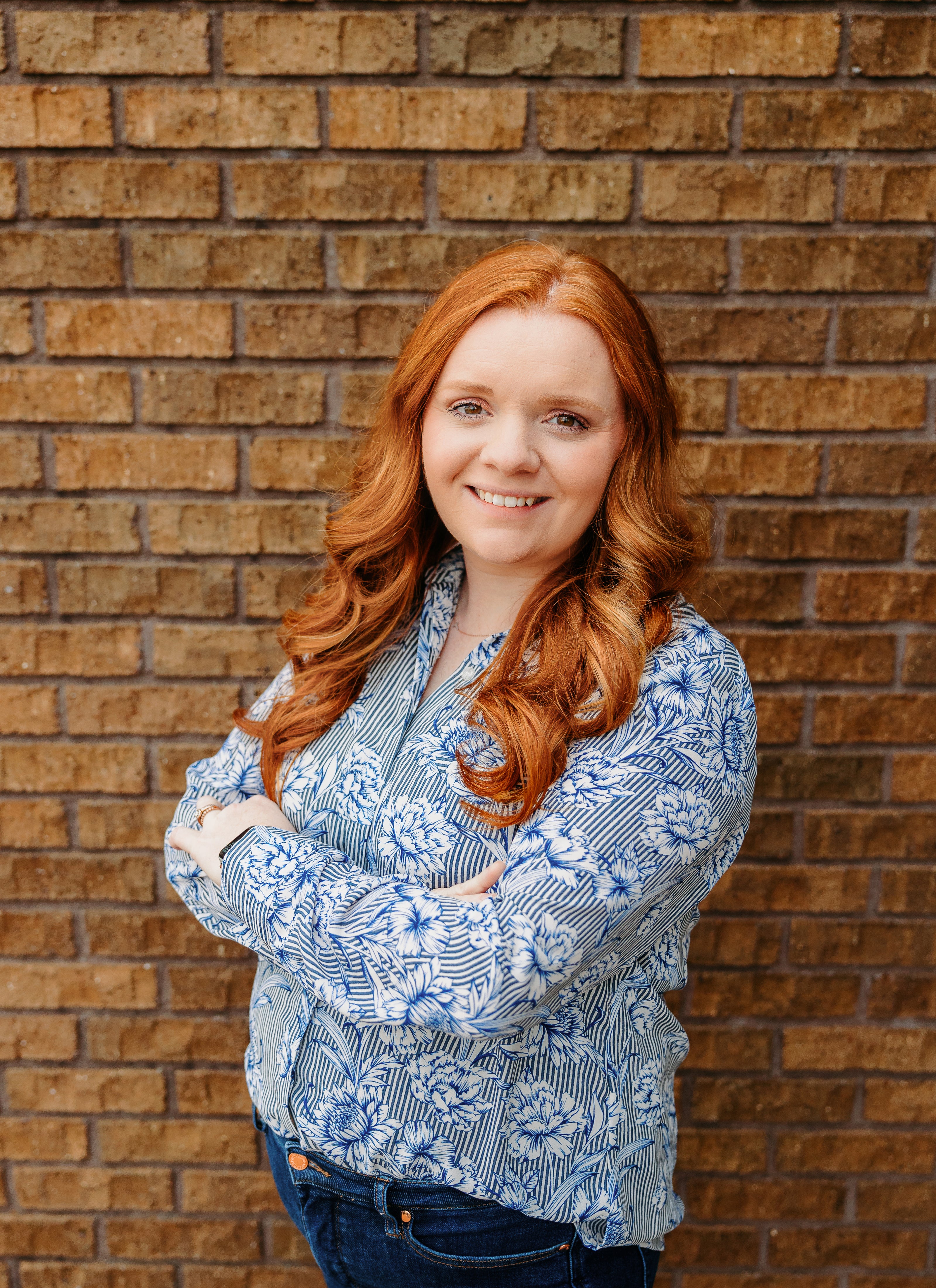 Woman with red hair wearing a blue patterned shirt against a brick wall.