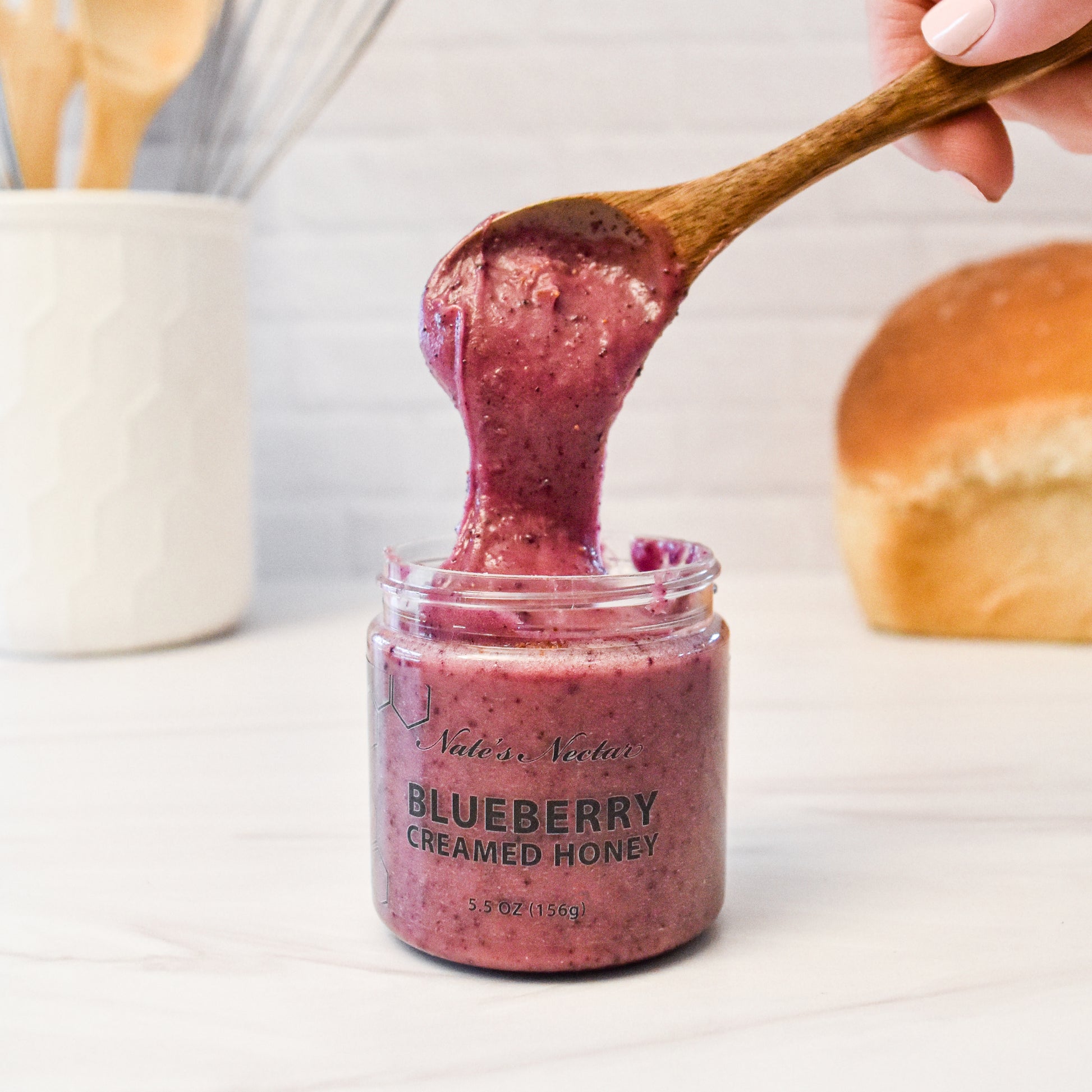 Jar of blueberry creamed honey with a wooden spoon lifting some out, on a white surface.