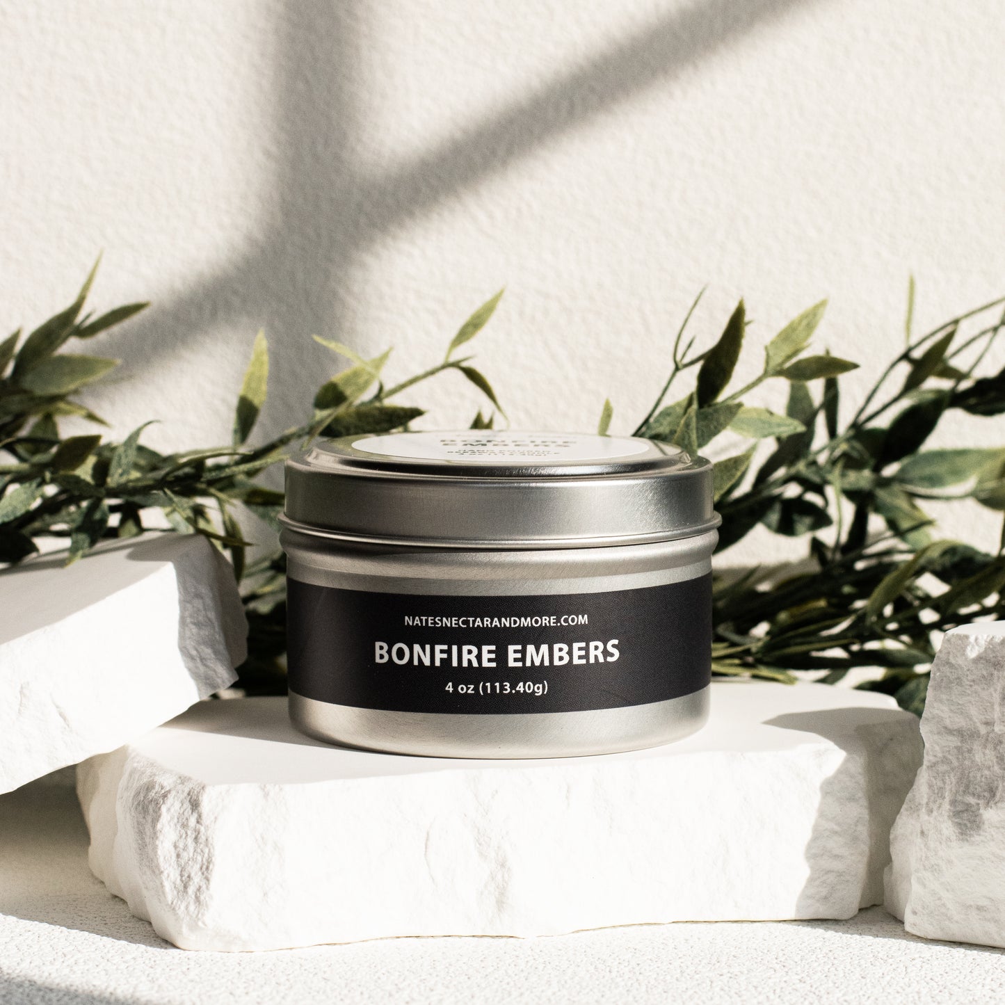 Candle tin labeled 'Bonfire Embers' on a white surface with greenery in the background