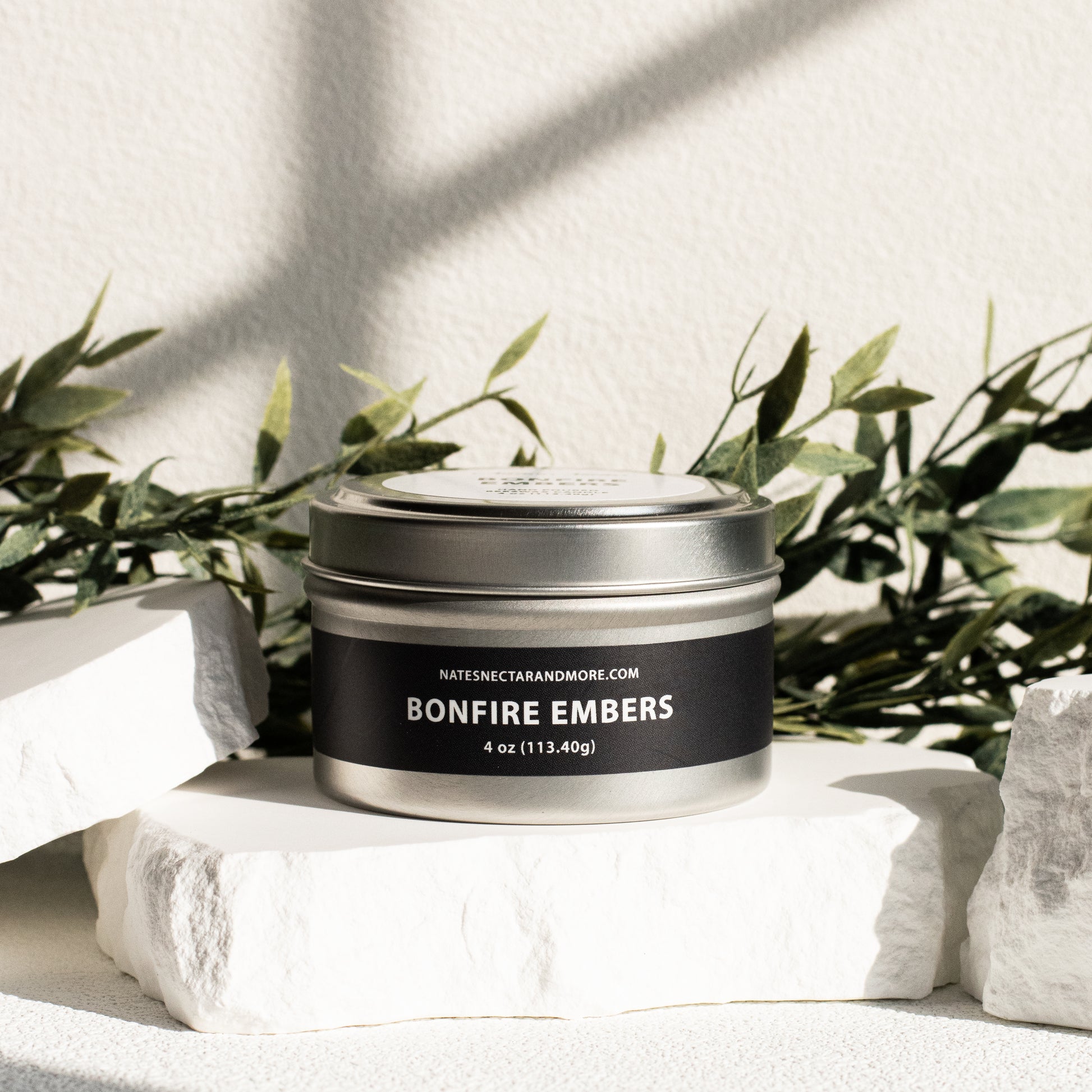 Candle tin labeled 'Bonfire Embers' on a white surface with greenery in the background