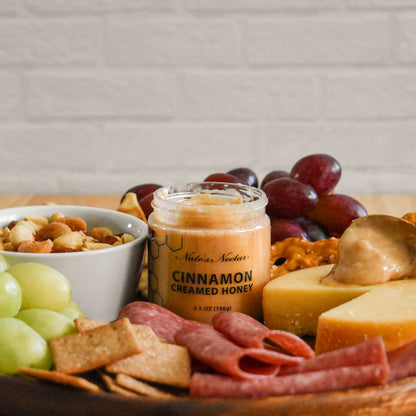 Jar of Natures Nectar cinnamon creamed honey on a charcuterie board with fruits, cheese, and nuts.