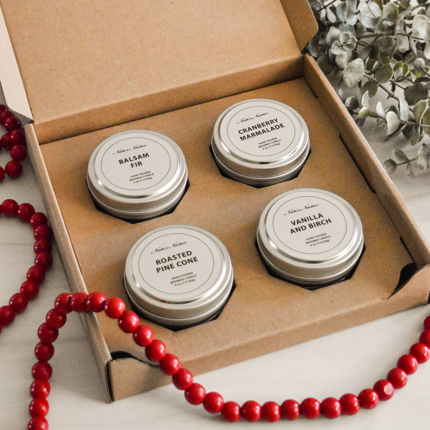 Four metal tins with labels in a cardboard box, surrounded by red beads and greenery.