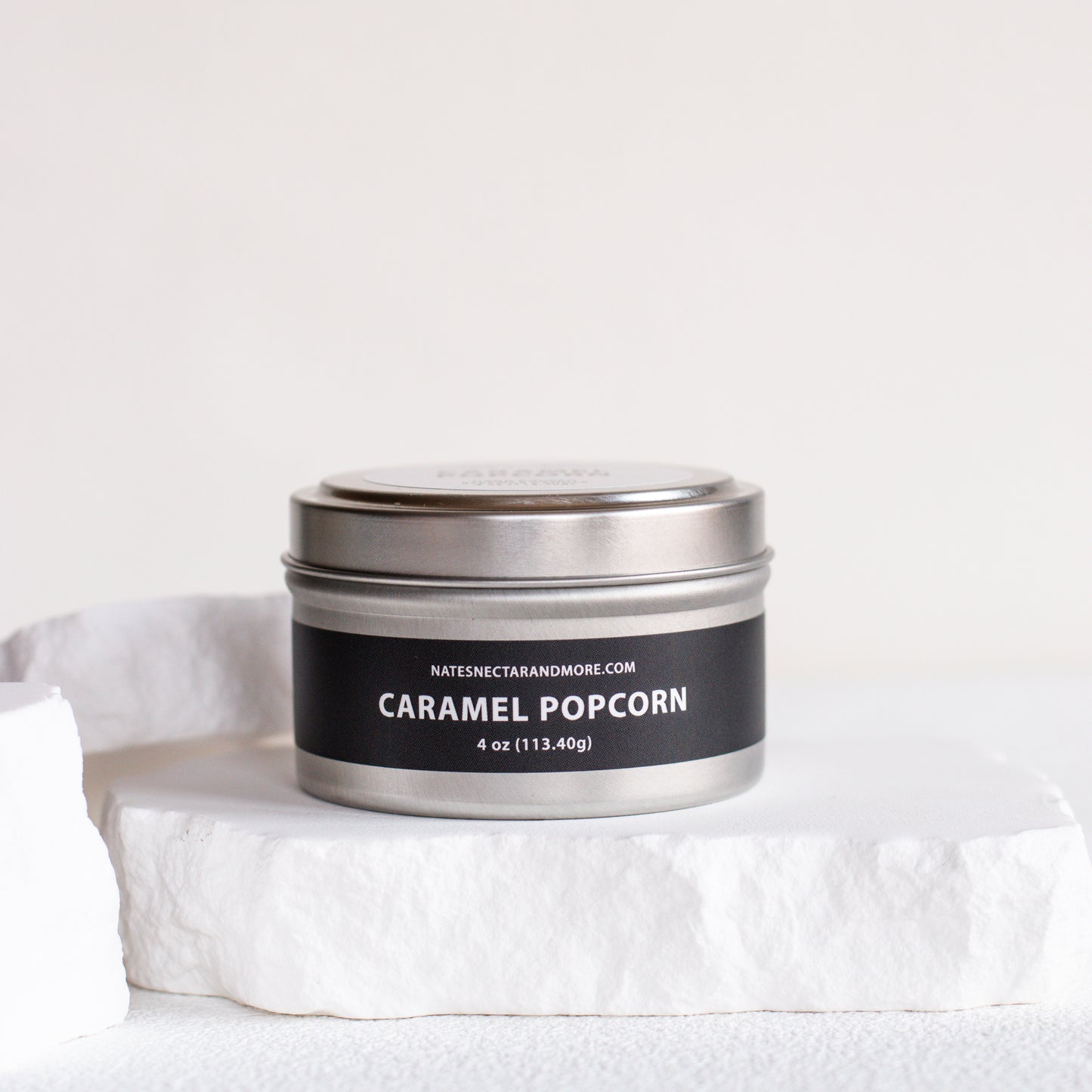 A silver metal tin candle labeled 'CARAMEL POPCORN' by Natescentarianmore.com, resting on a white stone pedestal against a clean white background.