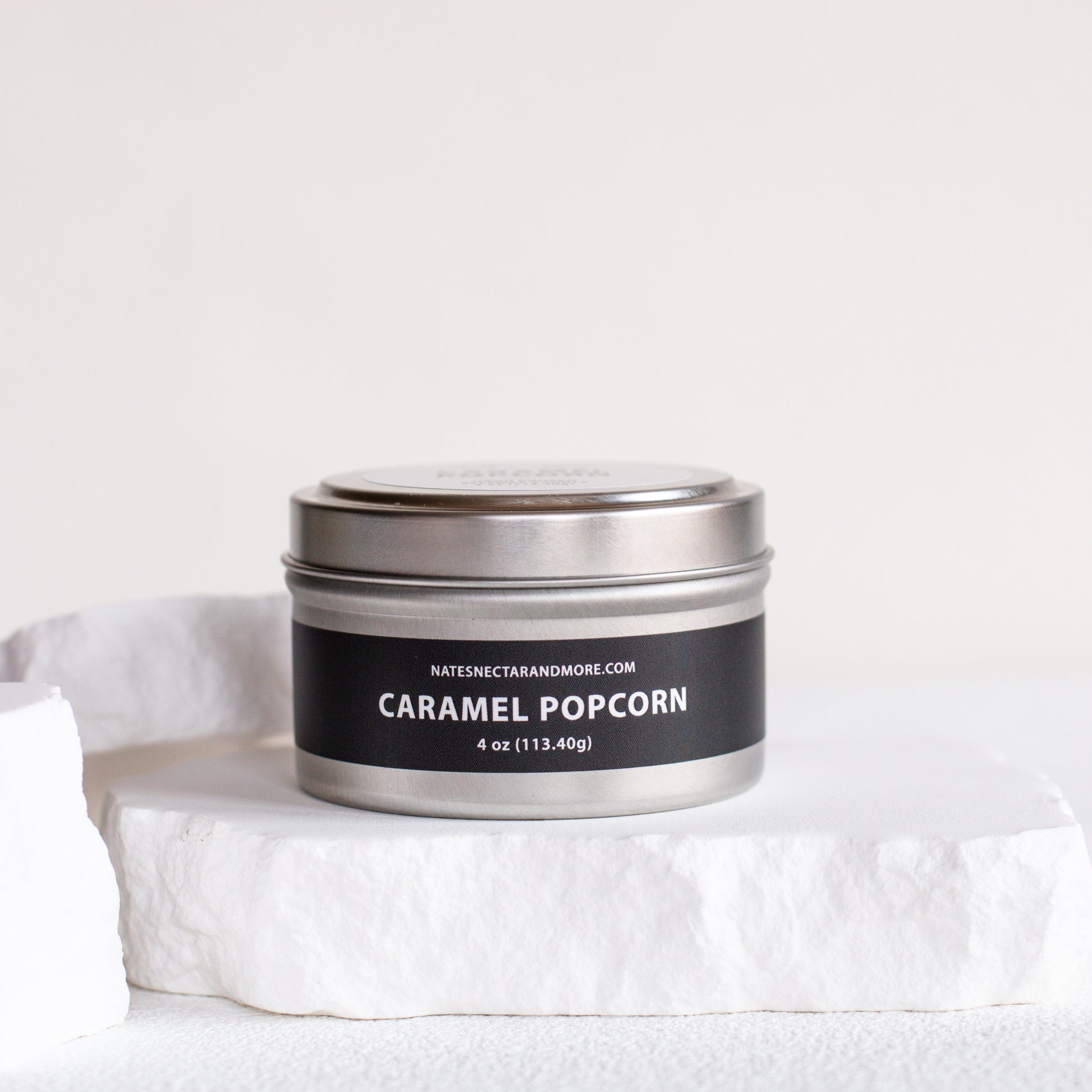 A silver metal tin candle labeled 'CARAMEL POPCORN' by Natescentarianmore.com, resting on a white stone pedestal against a clean white background.