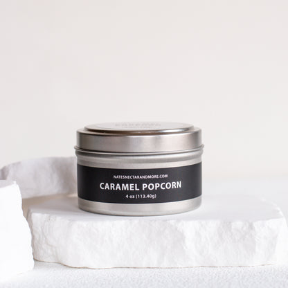 A silver metal tin candle labeled 'CARAMEL POPCORN' by Natescentarianmore.com, resting on a white stone pedestal against a clean white background.