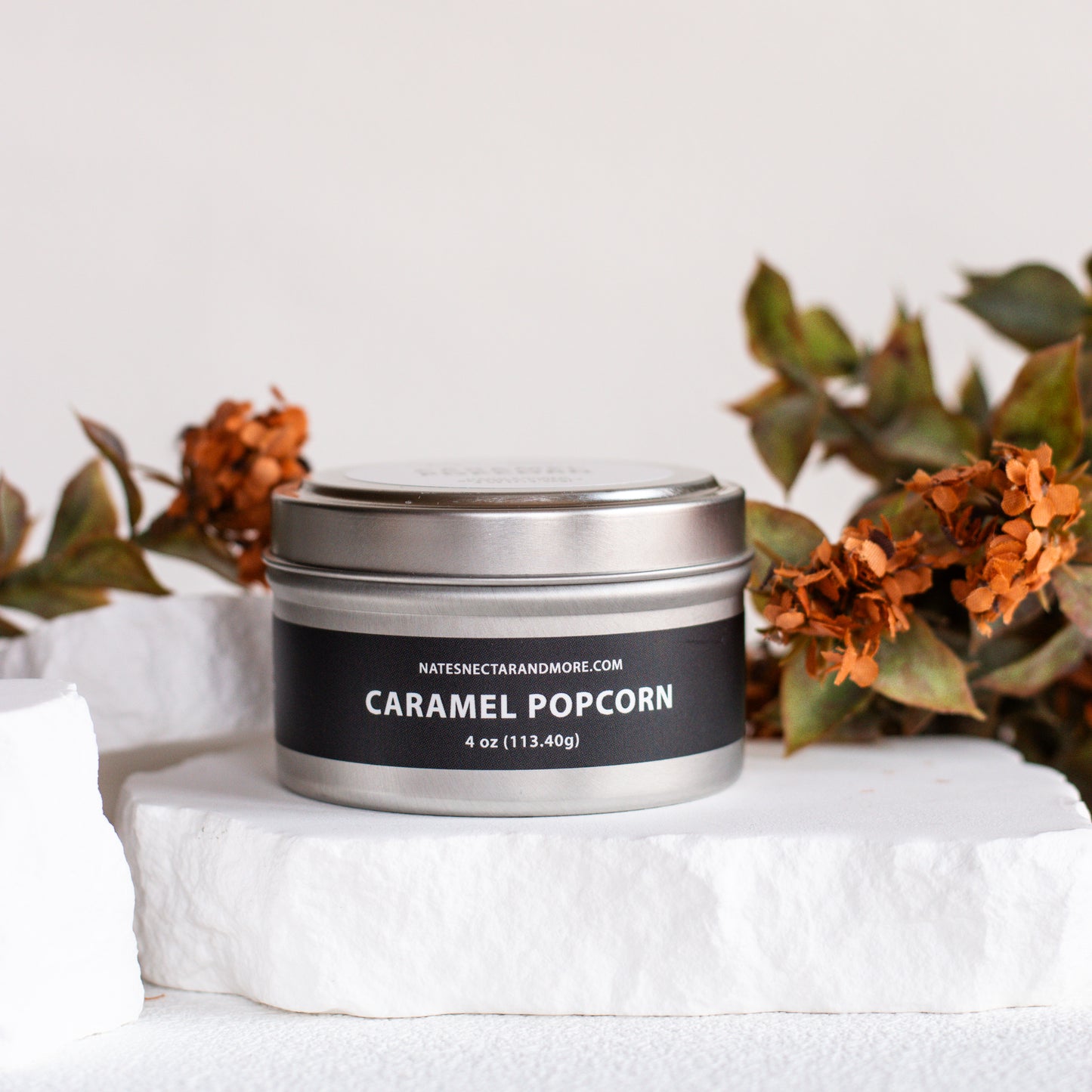 A silver metal tin candle labeled 'CARAMEL POPCORN' by Natescent Candles, displayed on a white stone pedestal with dried orange flowers in the background.