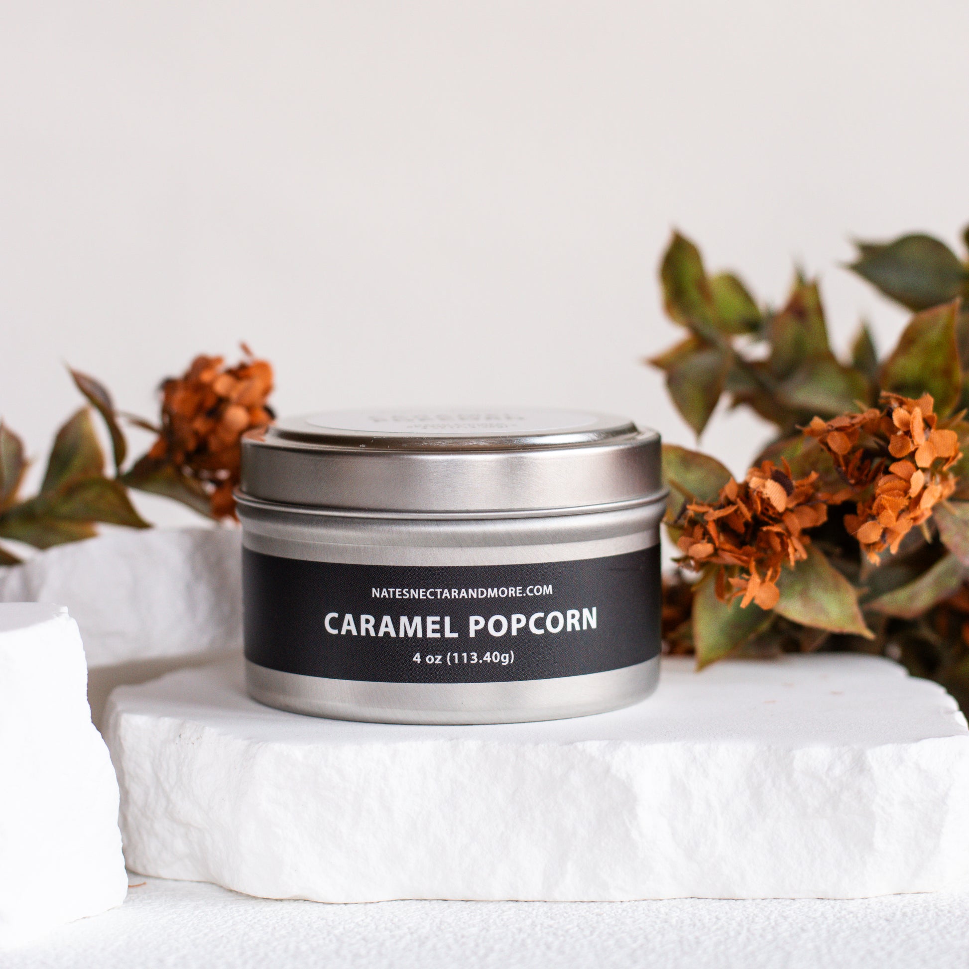 A silver metal tin candle labeled 'CARAMEL POPCORN' by Natescent Candles, displayed on a white stone pedestal with dried orange flowers in the background.