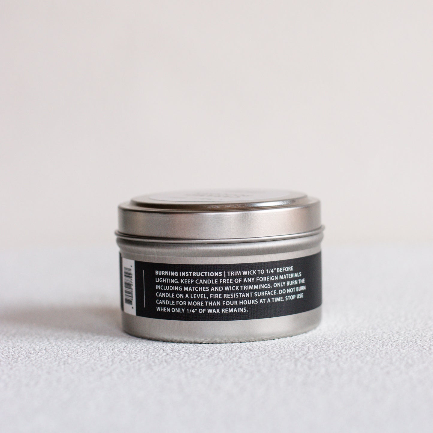 A small round metal tin candle shown from the back, with a black label containing white text and a barcode, set against a plain light background.