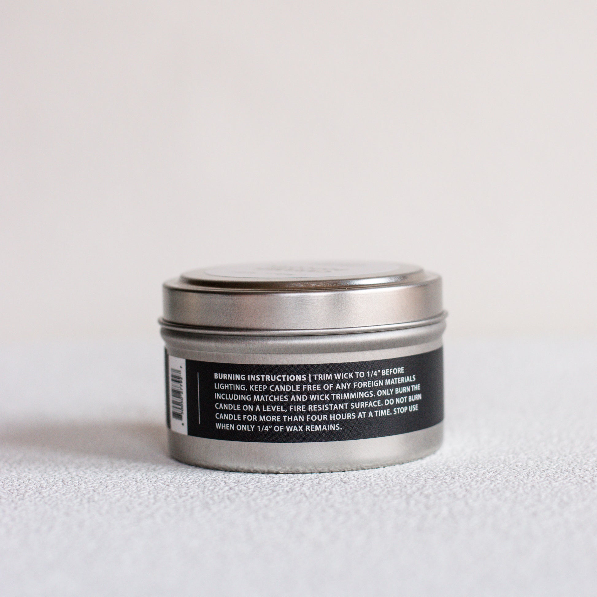 A small round metal tin candle shown from the back, with a black label containing white text and a barcode, set against a plain light background.