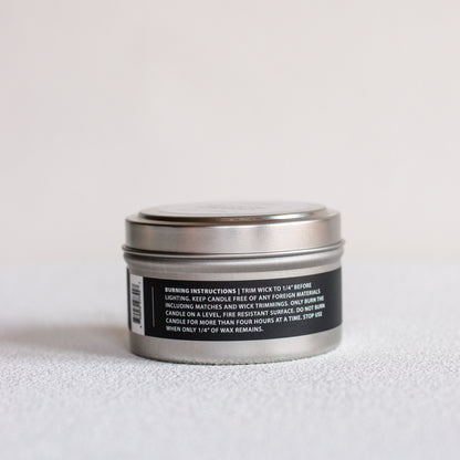 A small round metal tin candle shown from the back, with a black label containing white text and a barcode, set against a plain light background.
