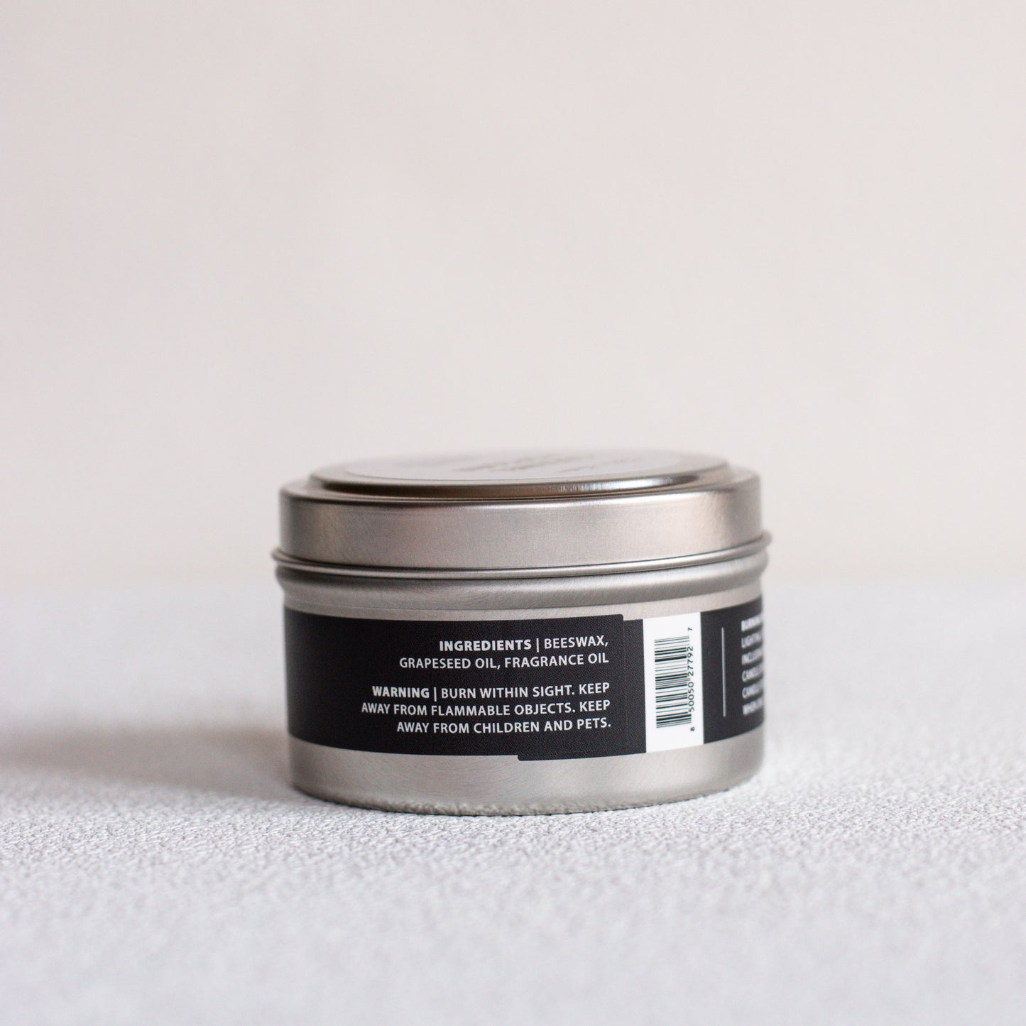 A small round metal tin candle with a black label, photographed on a light neutral surface against a soft light gray background.