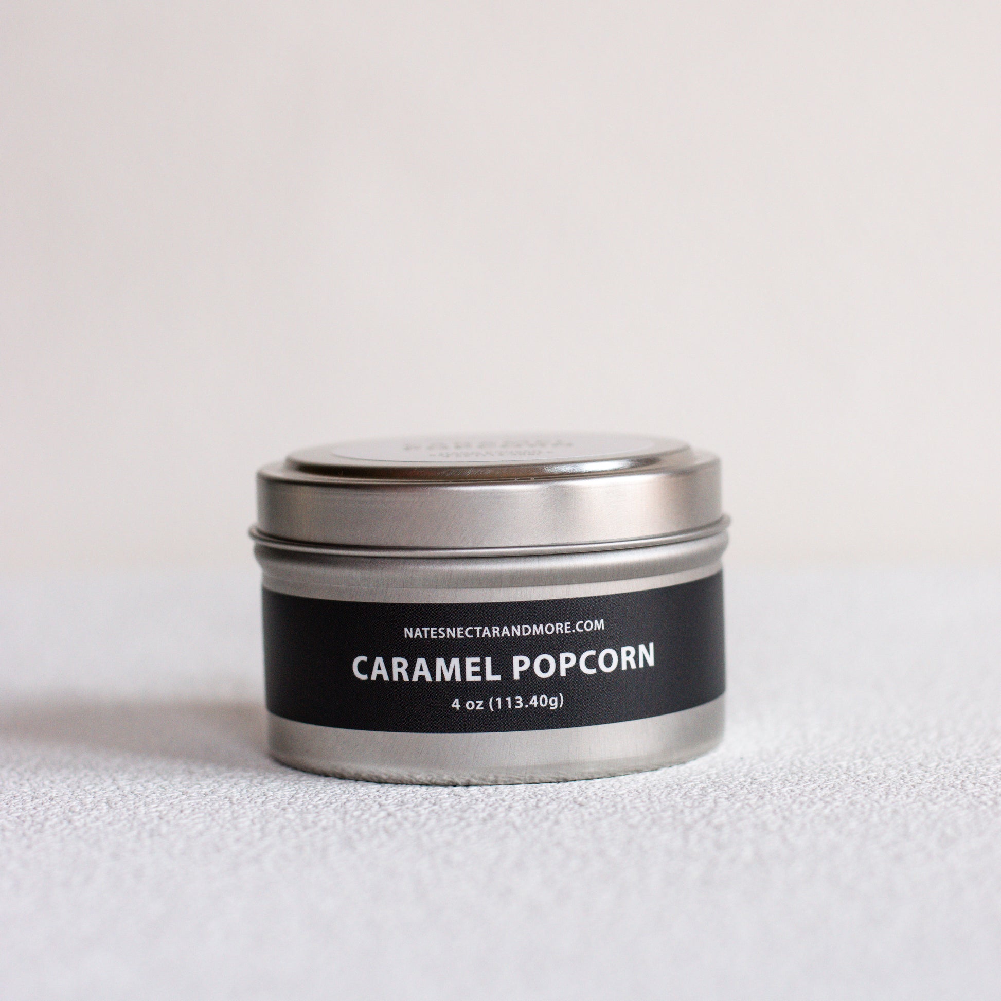 A small round metal tin candle labeled 'CARAMEL POPCORN' by Natescigarandmore.com, with a black label and silver lid, photographed on a light neutral surface.