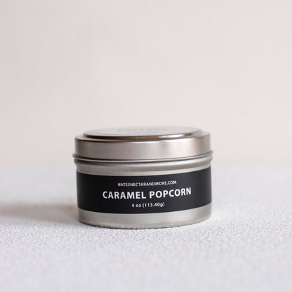 A small round metal tin candle labeled 'CARAMEL POPCORN' by Natescigarandmore.com, with a black label and silver lid, photographed on a light neutral surface.