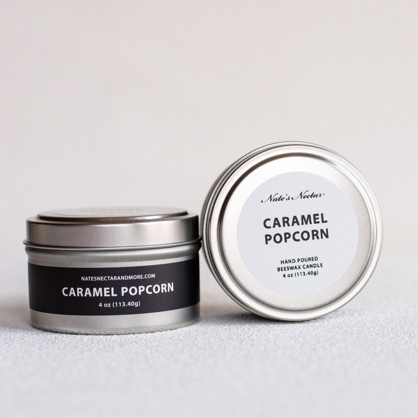 Two silver metal tins of Nates' Nectar Caramel Popcorn candles on a light gray textured surface. One tin is closed with a black label, and the other is open showing the white label inside.