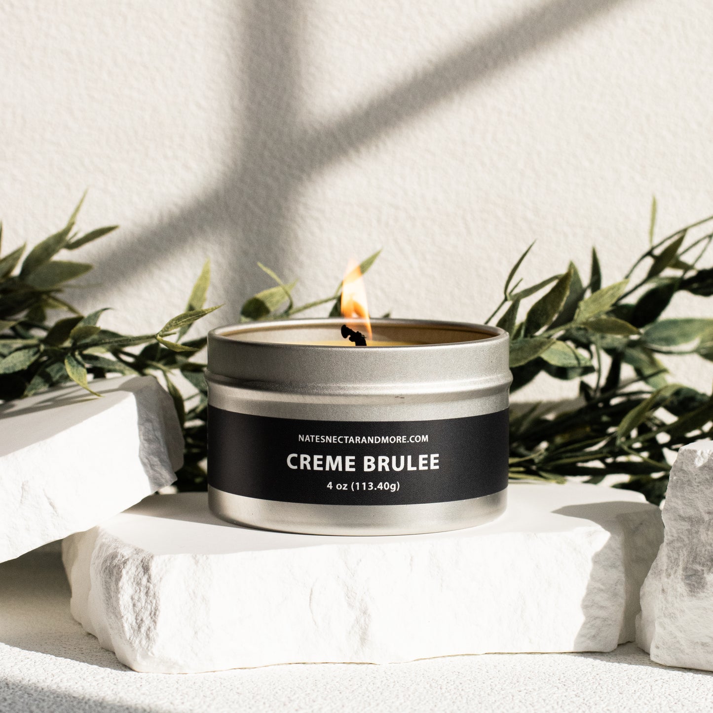 Candle in a tin labeled 'Creme Brulee' with greenery in the background