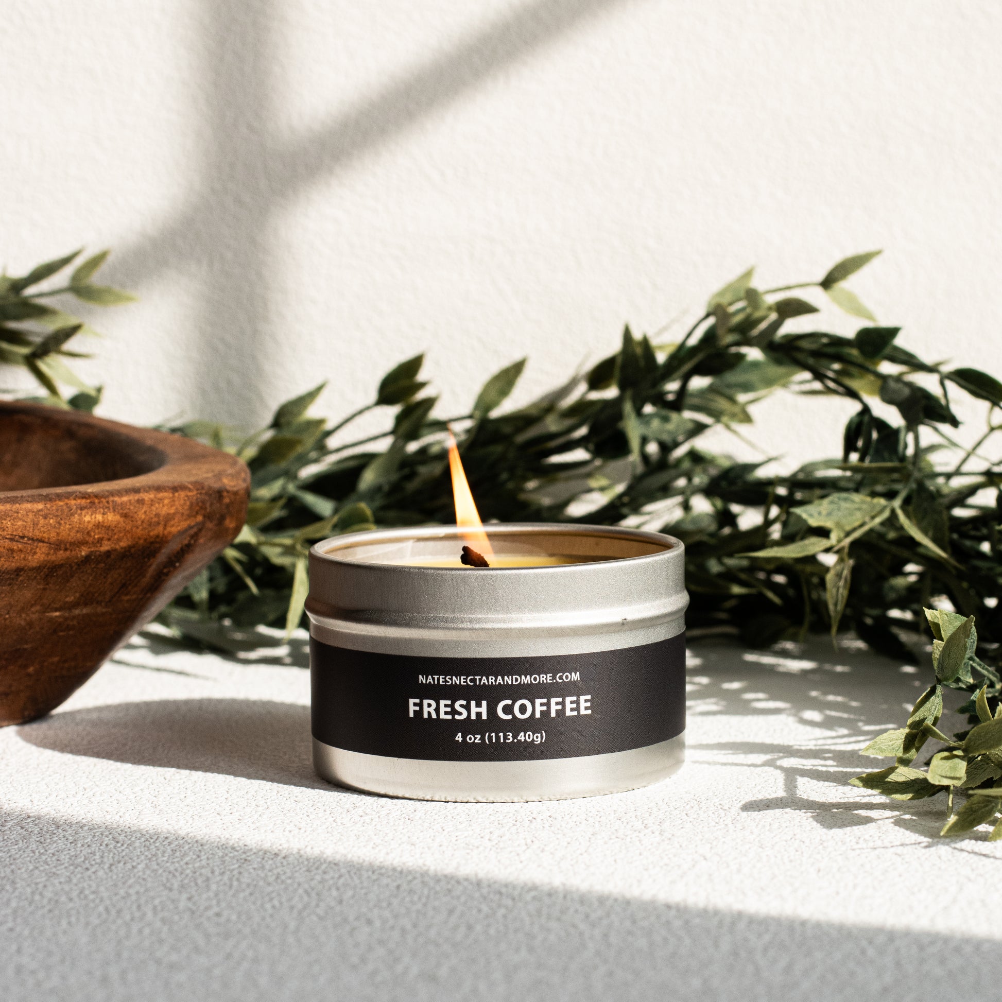 Candle labeled 'Fresh Coffee' in a metallic tin with a wooden bowl and greenery in the background.