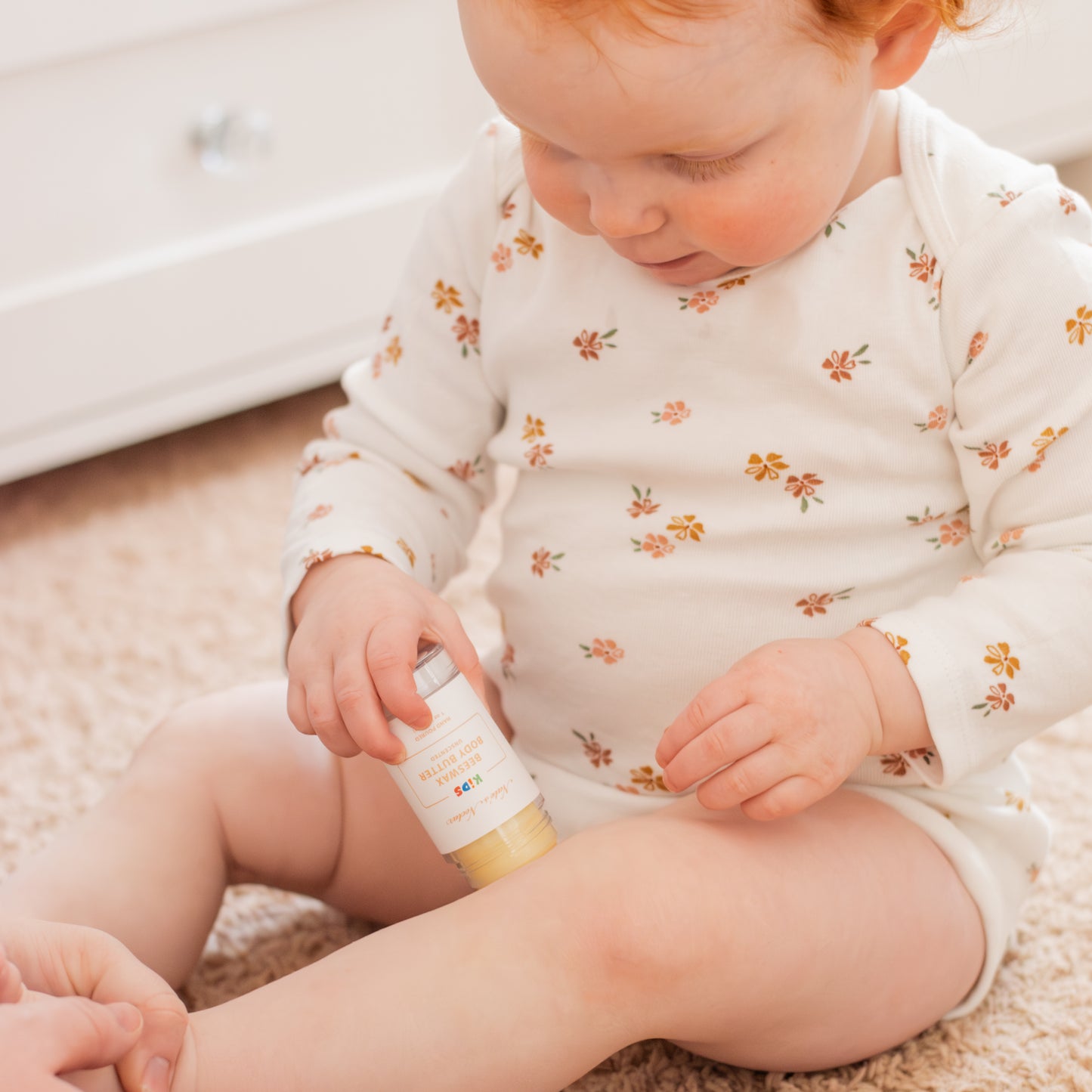 Baby in a floral onesie applying kids beeswax body butter to leg