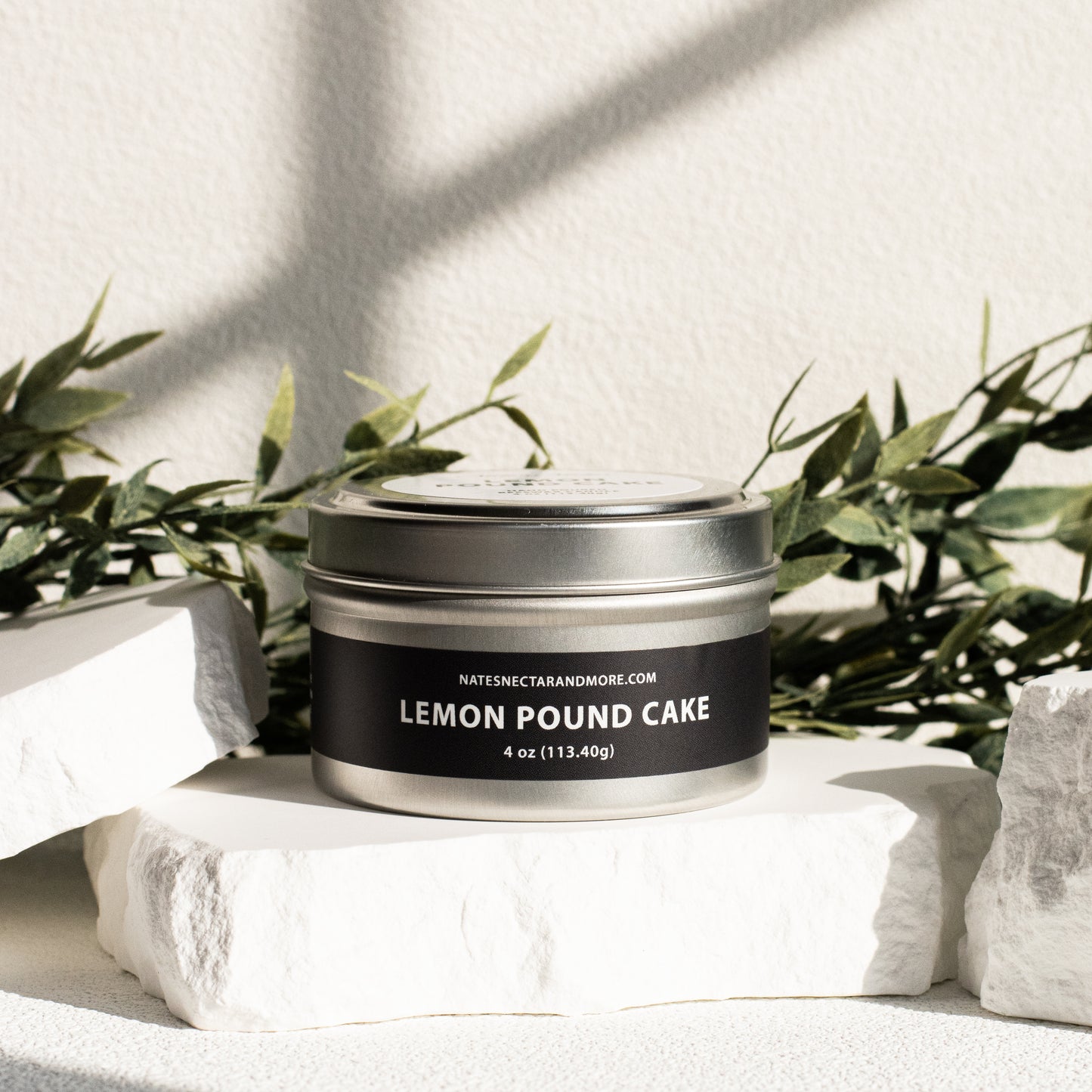 Candle in a tin labeled 'Lemon Pound Cake' on a white surface with greenery in the background