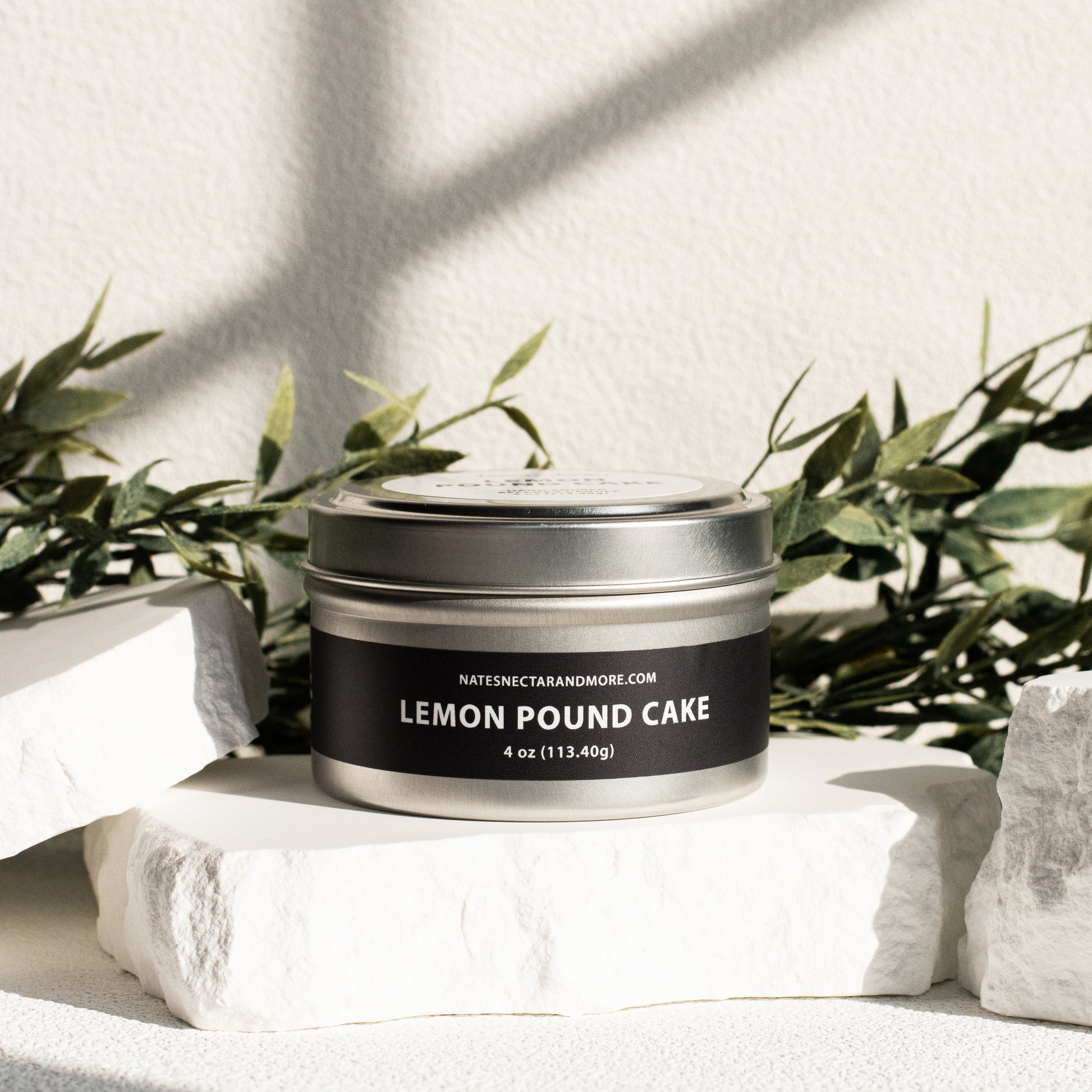 Candle in a tin labeled 'Lemon Pound Cake' on a white surface with greenery in the background