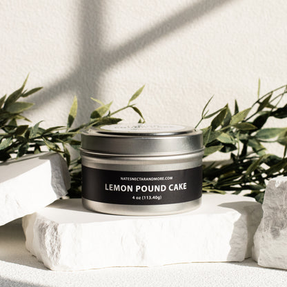 Candle in a tin labeled 'Lemon Pound Cake' on a white surface with greenery in the background
