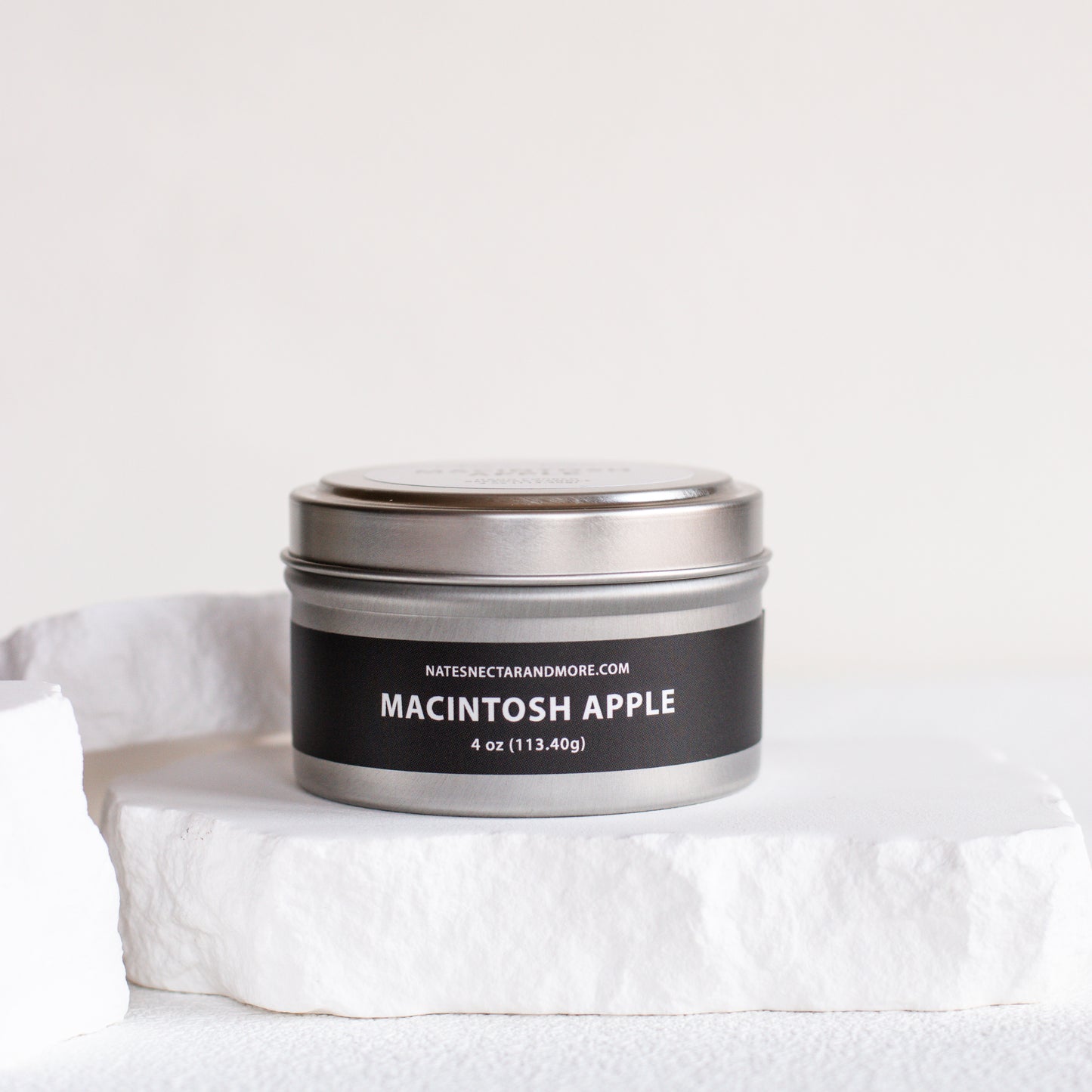 A 4 oz metal tin candle labeled 'MACINTOSH APPLE' by Nature's Craft and More, sitting on a white stone pedestal against a clean white background.