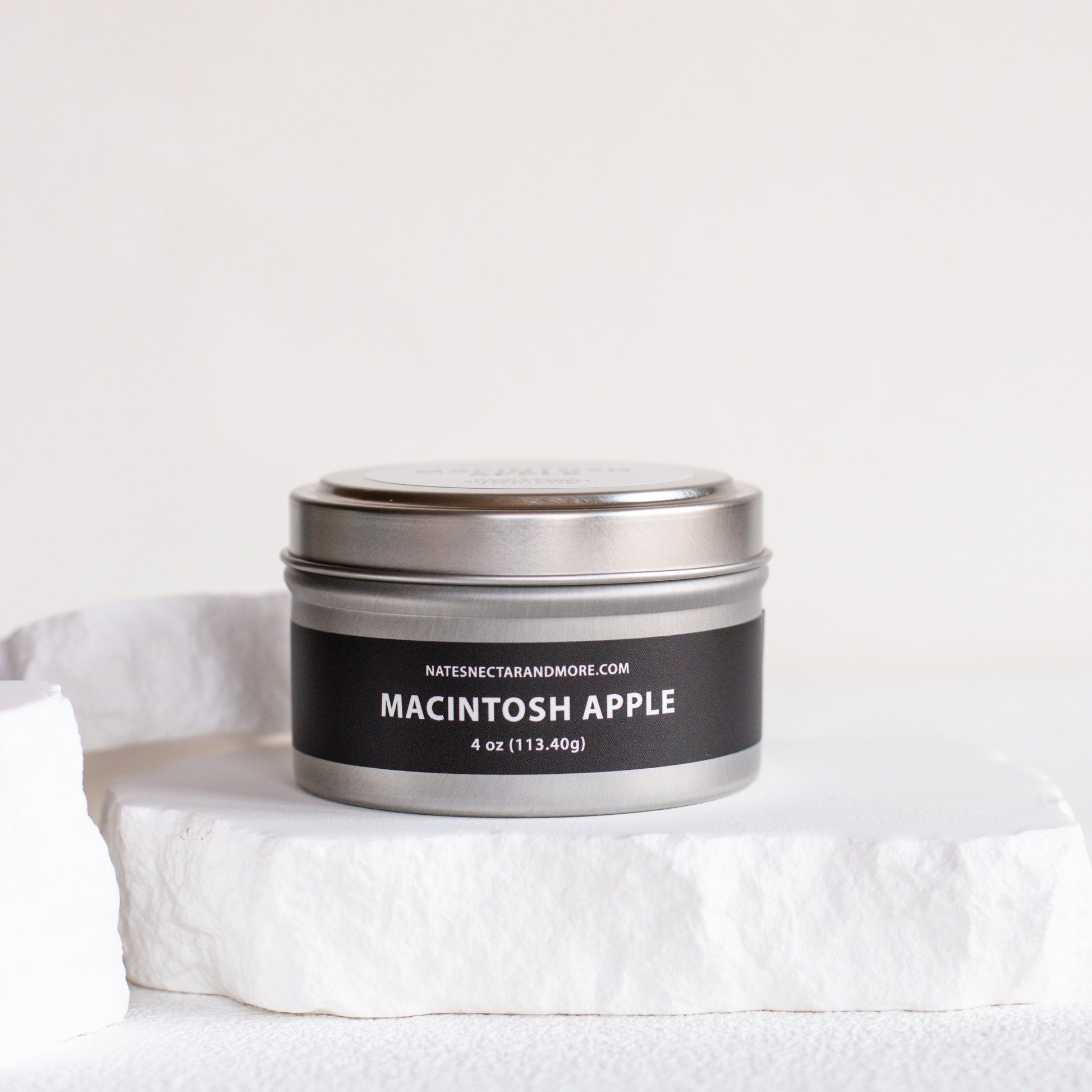 A 4 oz metal tin candle labeled 'MACINTOSH APPLE' by Nature's Craft and More, sitting on a white stone pedestal against a clean white background.