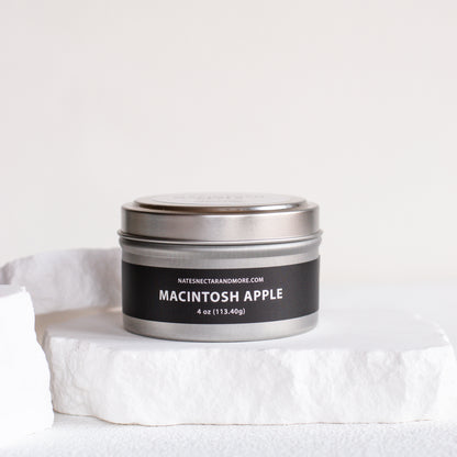 A 4 oz metal tin candle labeled 'MACINTOSH APPLE' by Nature's Craft and More, sitting on a white stone pedestal against a clean white background.