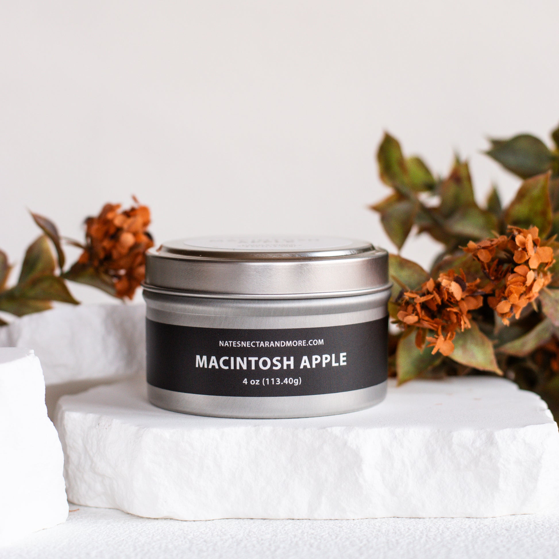 A silver metal tin candle labeled 'MACINTOSH APPLE' by Nature's Candle Company, styled on a white stone slab with dried orange flowers in the background.
