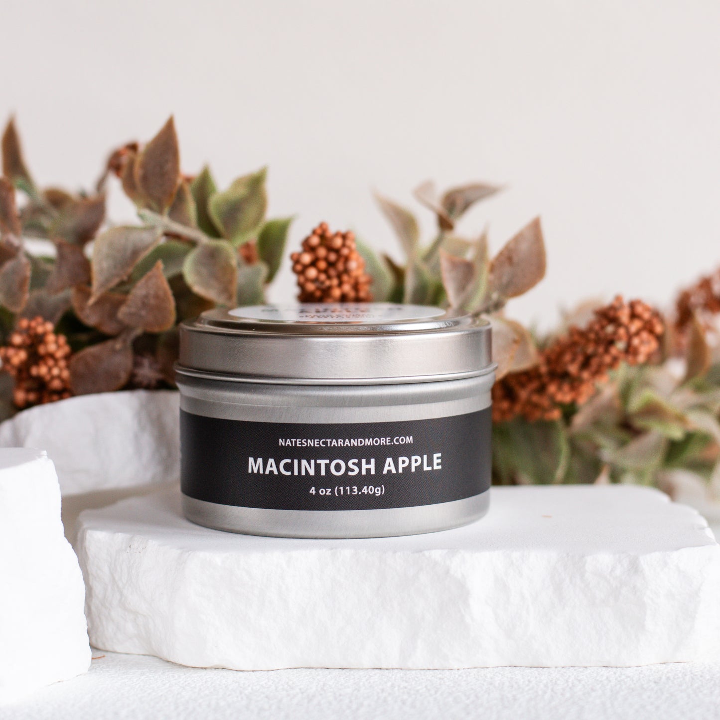A silver tin candle labeled 'MACINTOSH APPLE' by Nature's Secret, displayed on a white stone slab with dried botanicals in the background.