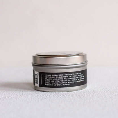 A small round metal tin candle with a silver lid and a black label showing burning instructions, photographed on a light gray textured surface against a white background.