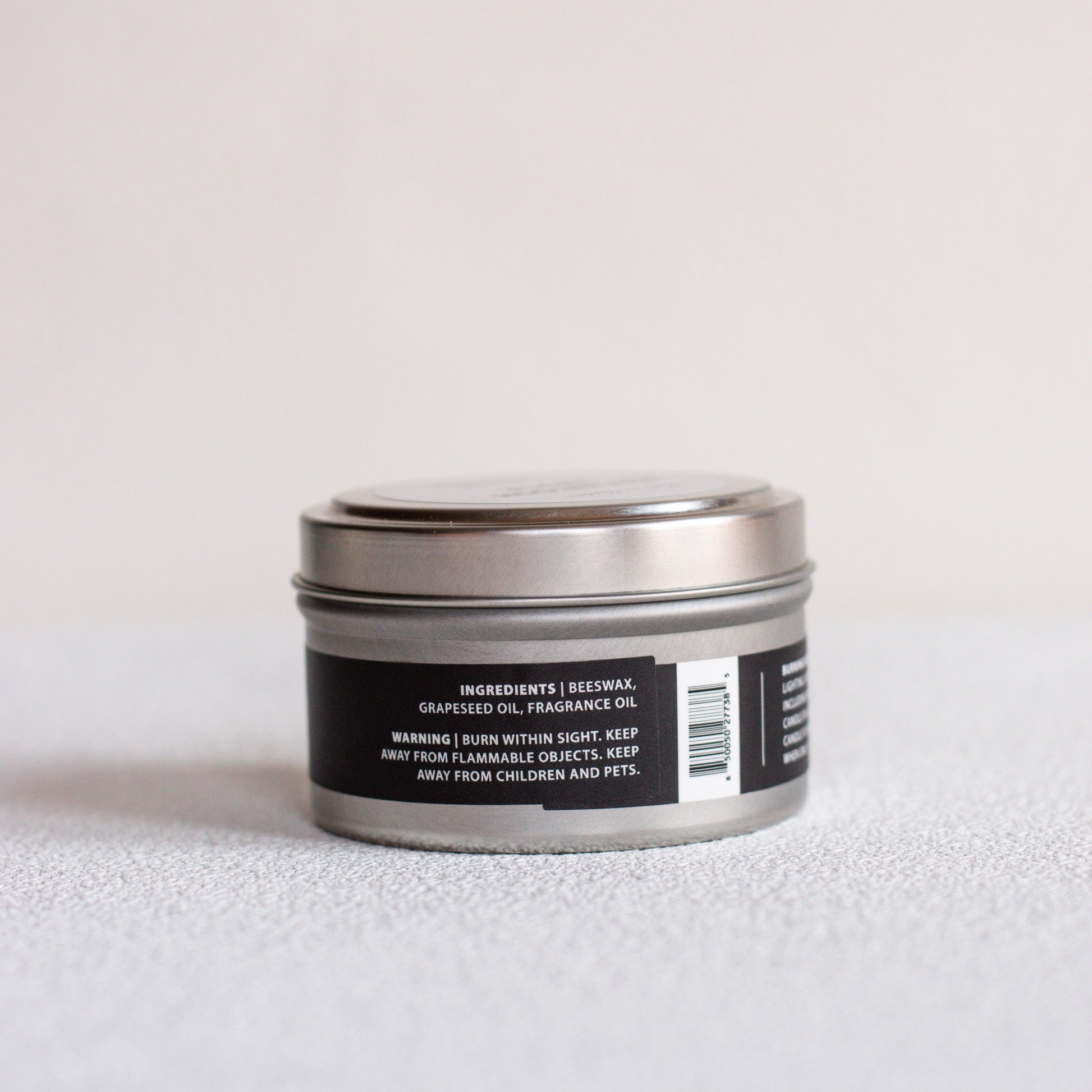 A small round metal tin candle with a black label, photographed on a light gray textured surface against a white background.