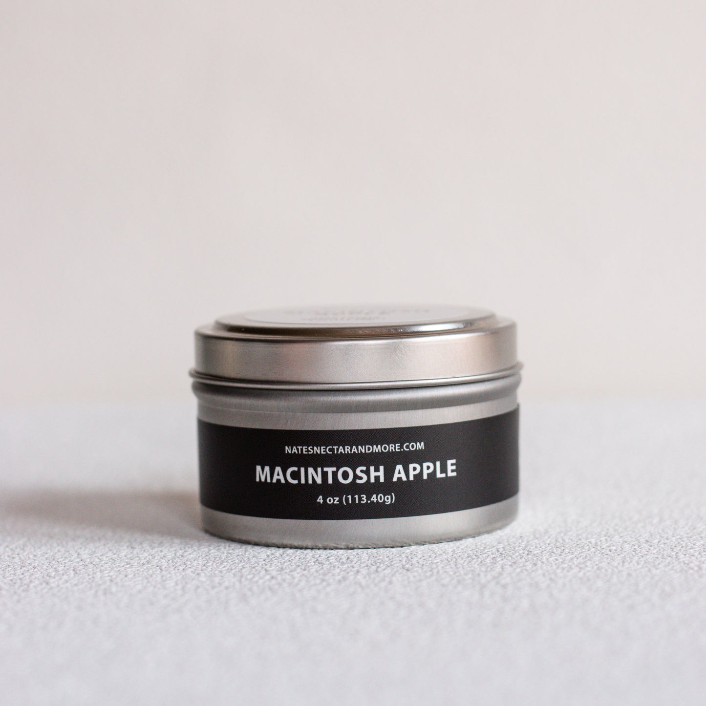 A small round metal tin candle labeled 'MACINTOSH APPLE' by Nates Nectar and More, with a black label and silver lid, photographed on a light neutral surface against a pale background.