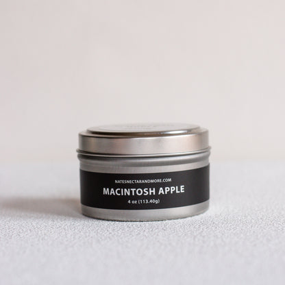 A small round metal tin candle labeled 'MACINTOSH APPLE' by Nates Nectar and More, with a black label and silver lid, photographed on a light neutral surface against a pale background.