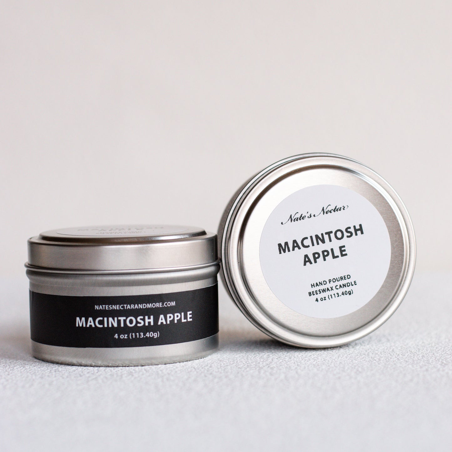 Two silver metal tin candles on a light gray surface, one closed with a black label reading Macintosh Apple and the other open showing the white lid label.