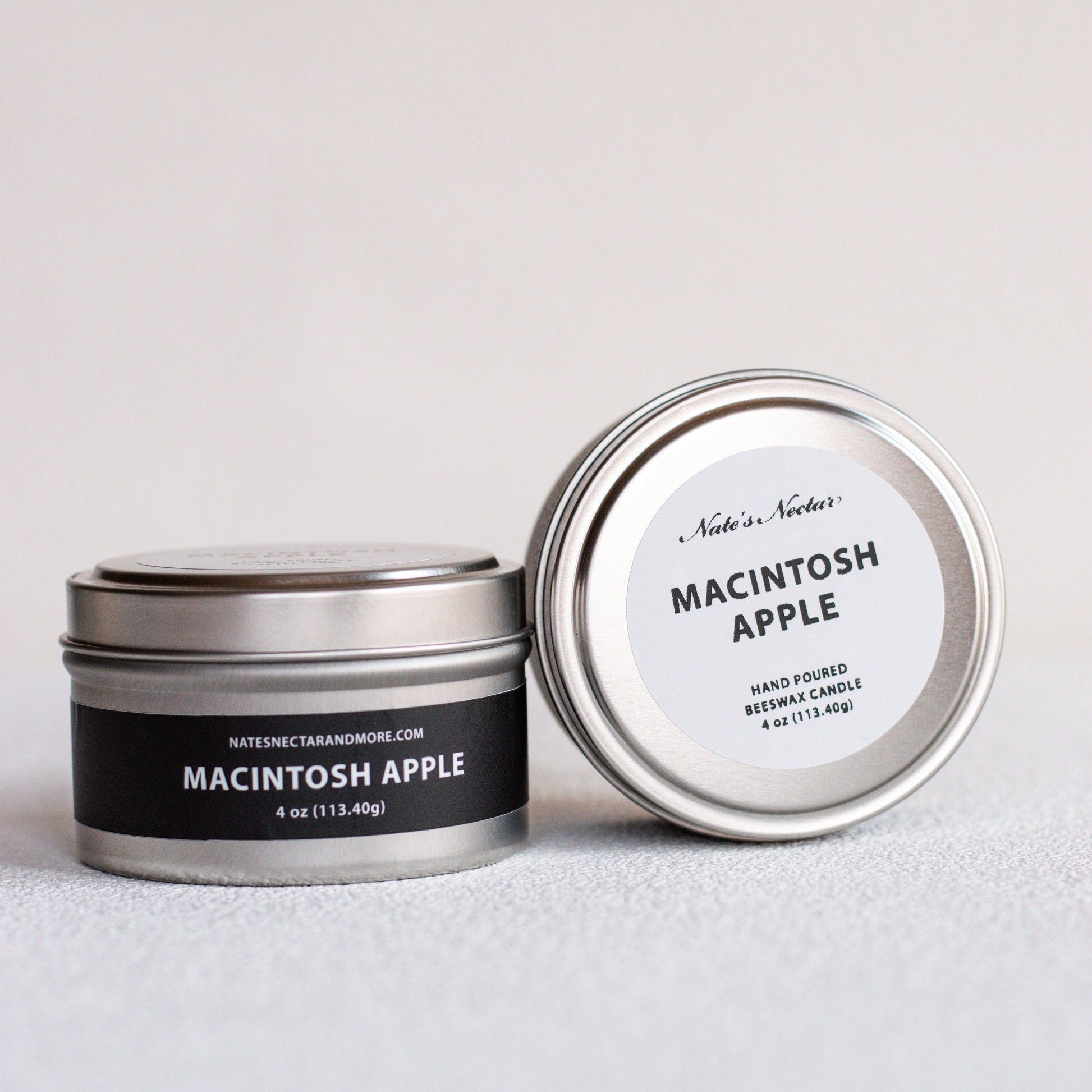 Two silver metal tin candles on a light gray surface, one closed with a black label reading Macintosh Apple and the other open showing the white lid label.