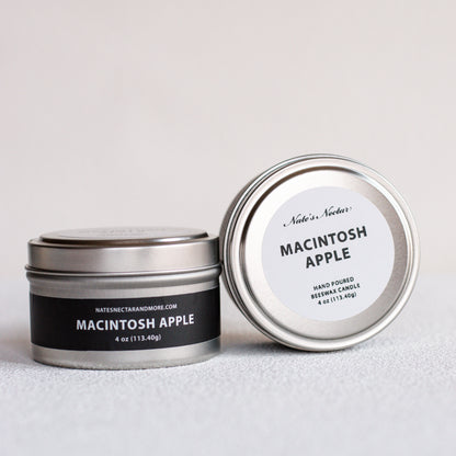Two silver metal tin candles on a light gray surface, one closed with a black label reading Macintosh Apple and the other open showing the white lid label.