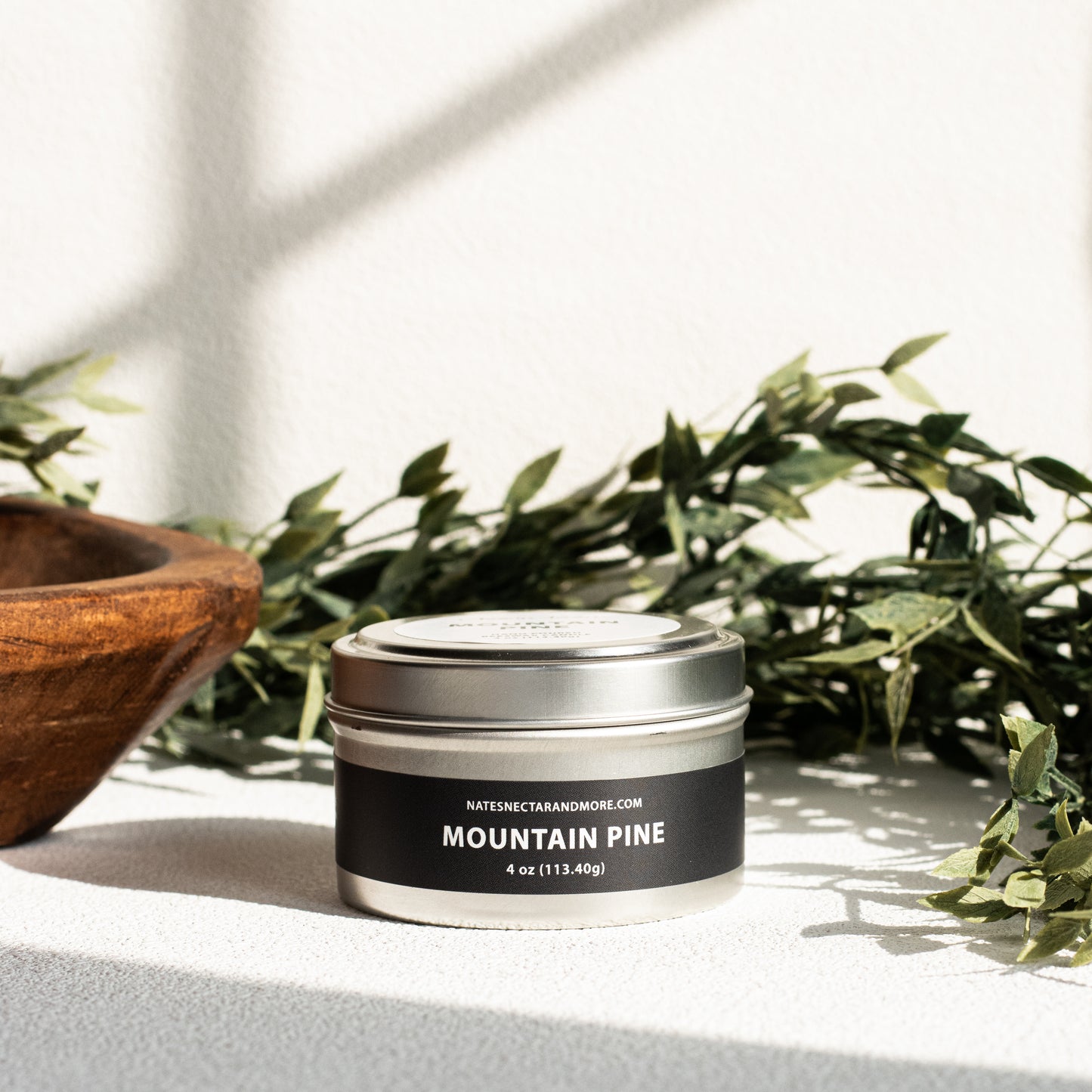 Candle in a metal tin labeled 'Mountain Pine' on a surface with greenery in the background
