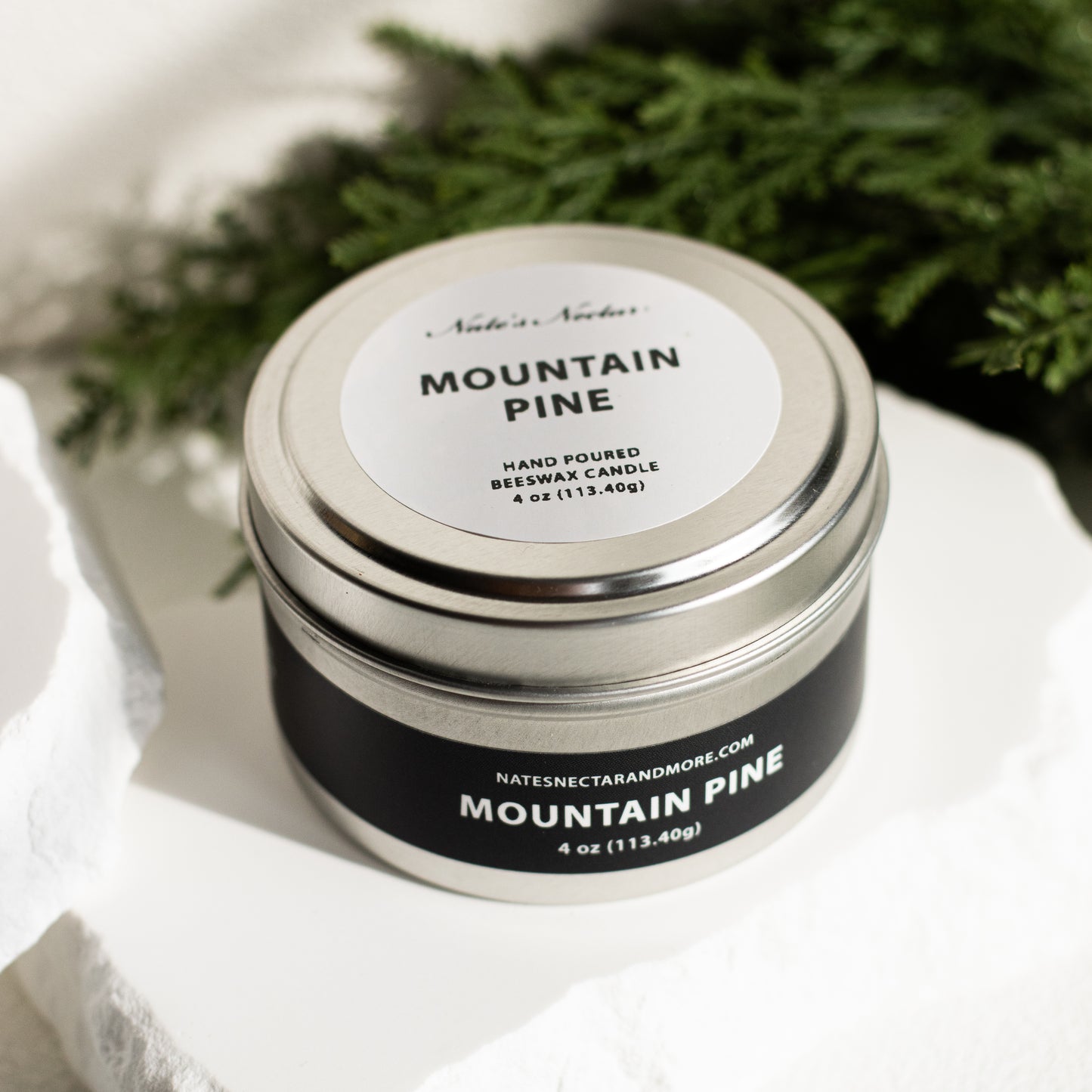 Candle jar labeled 'Mountain Pine' on a white surface with greenery in the background
