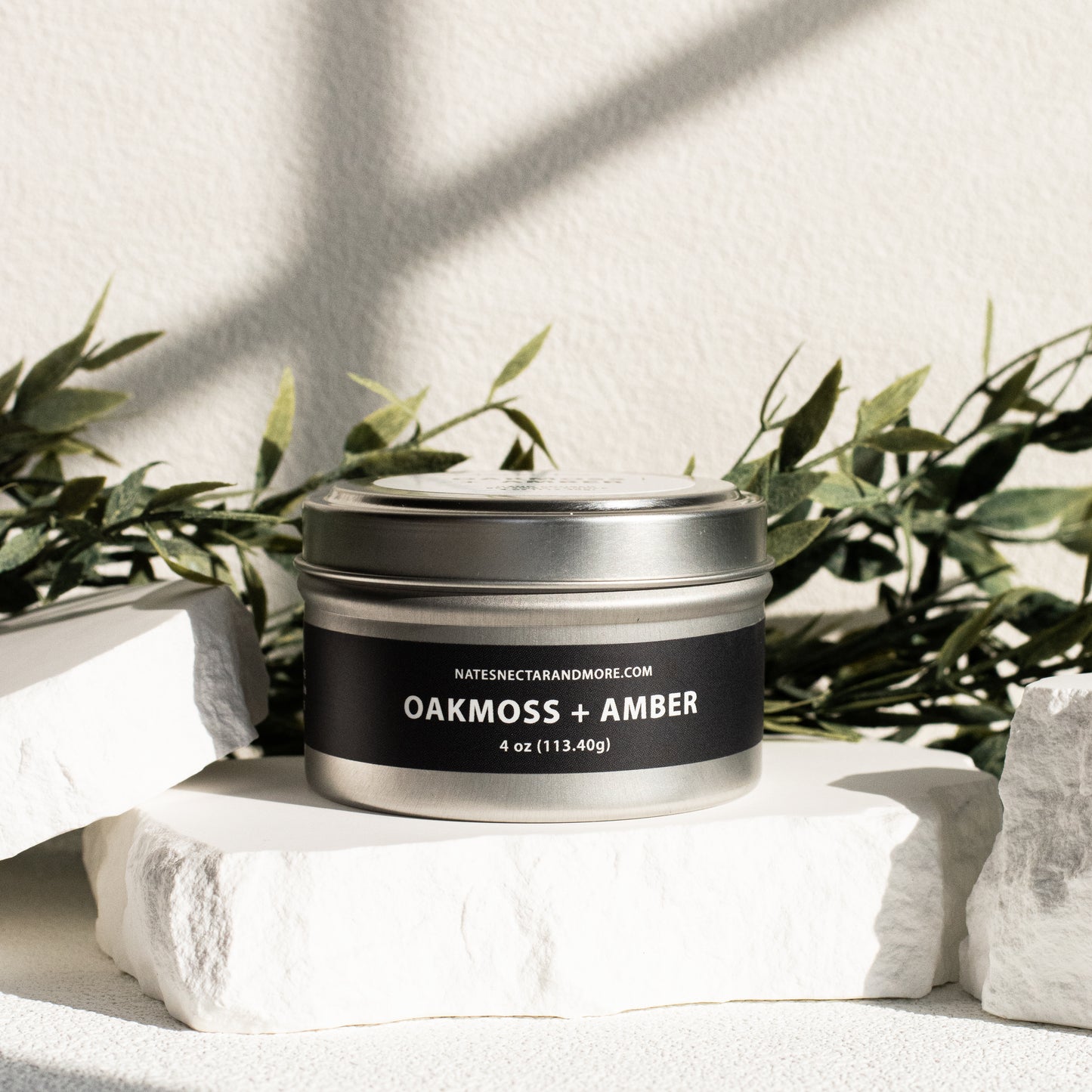 Candle jar labeled 'Oakmoss + Amber' on a white surface with greenery in the background