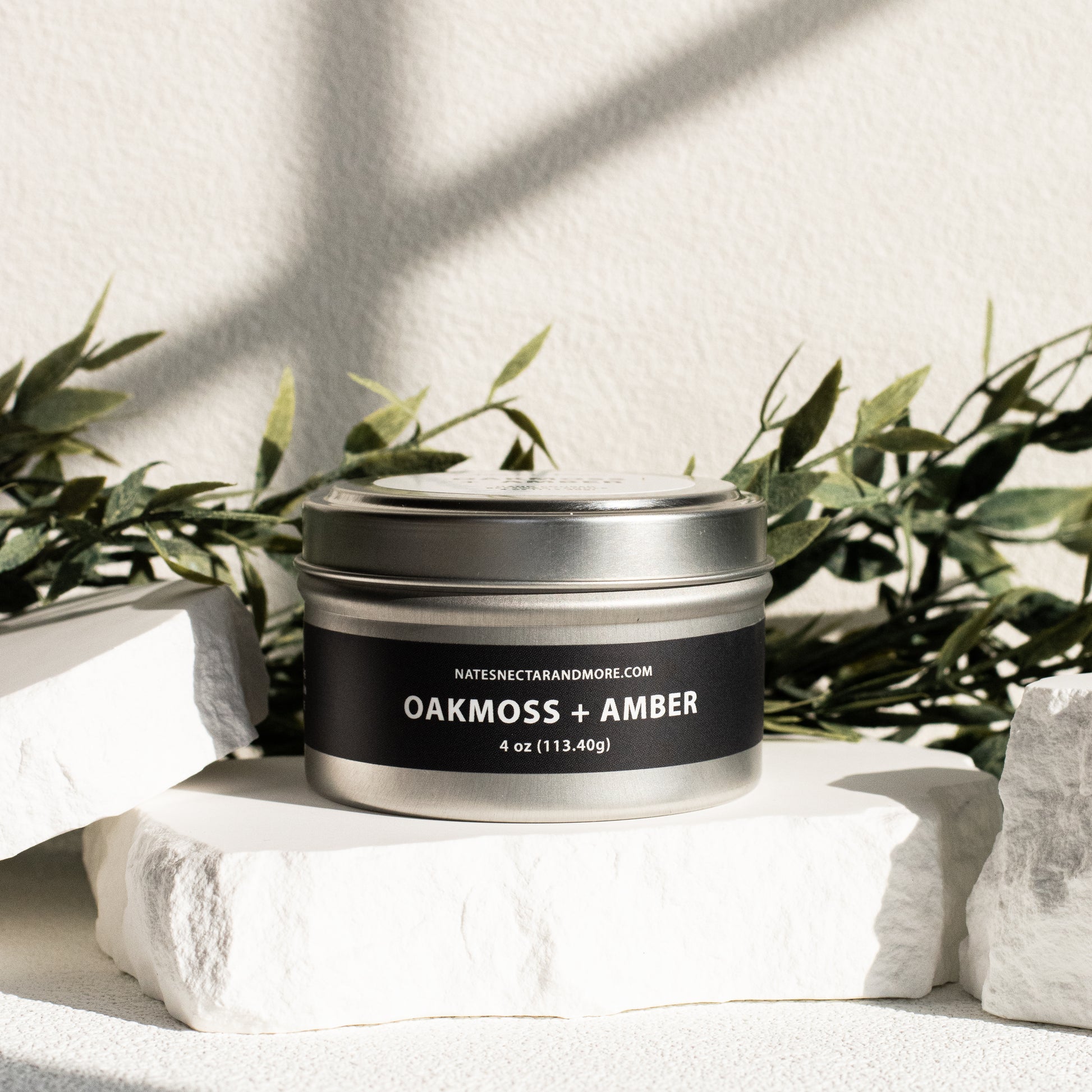 Candle jar labeled 'Oakmoss + Amber' on a white surface with greenery in the background