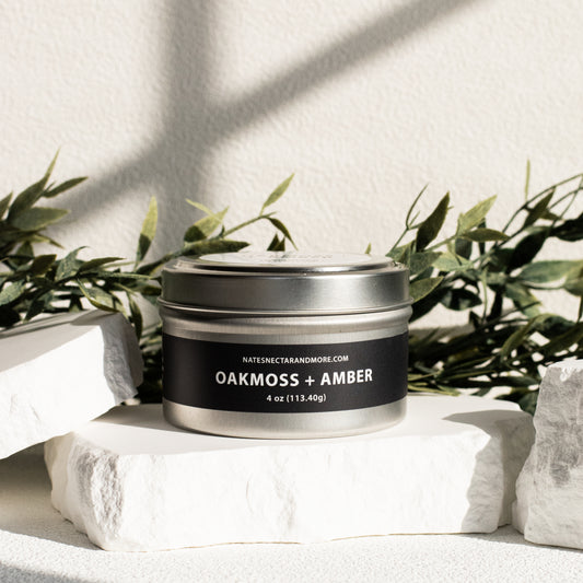 Candle jar labeled 'Oakmoss + Amber' on a white surface with greenery in the background