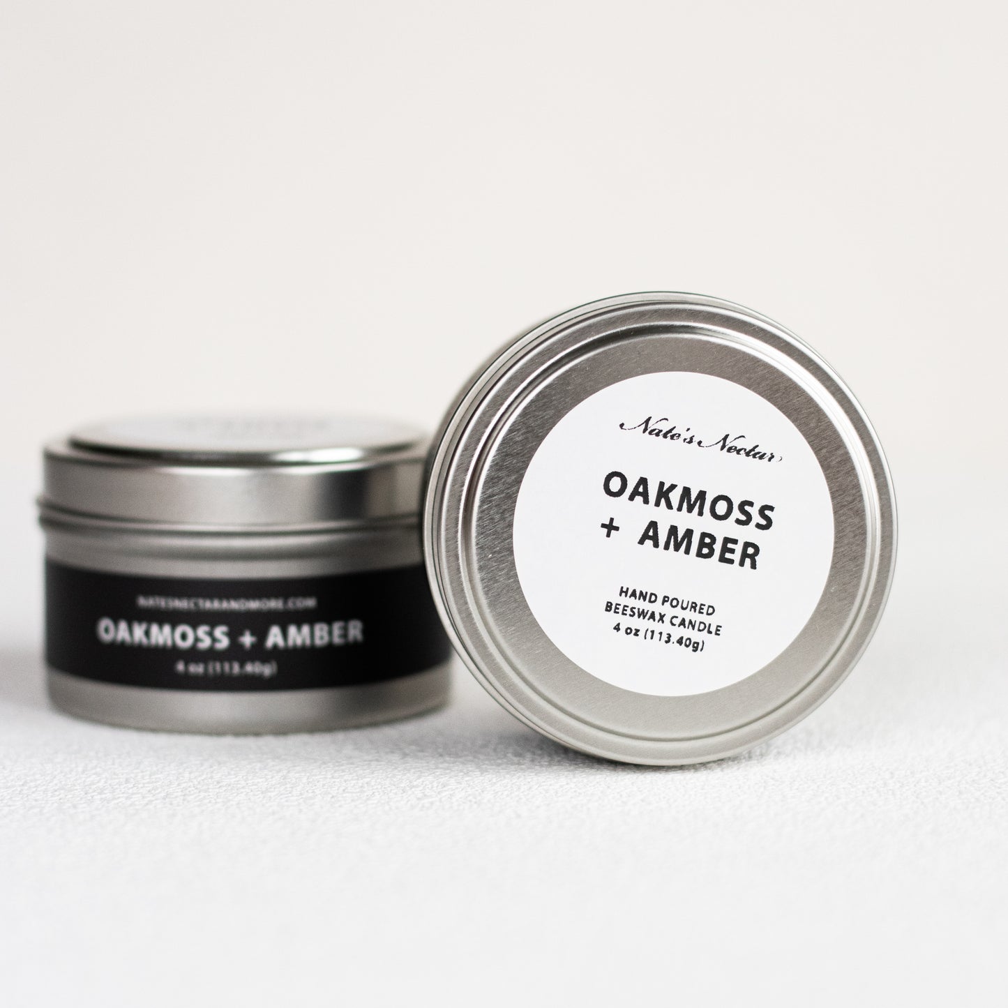 Two tins of 'Oakmoss + Amber' candles by Nade's Nook on a white background