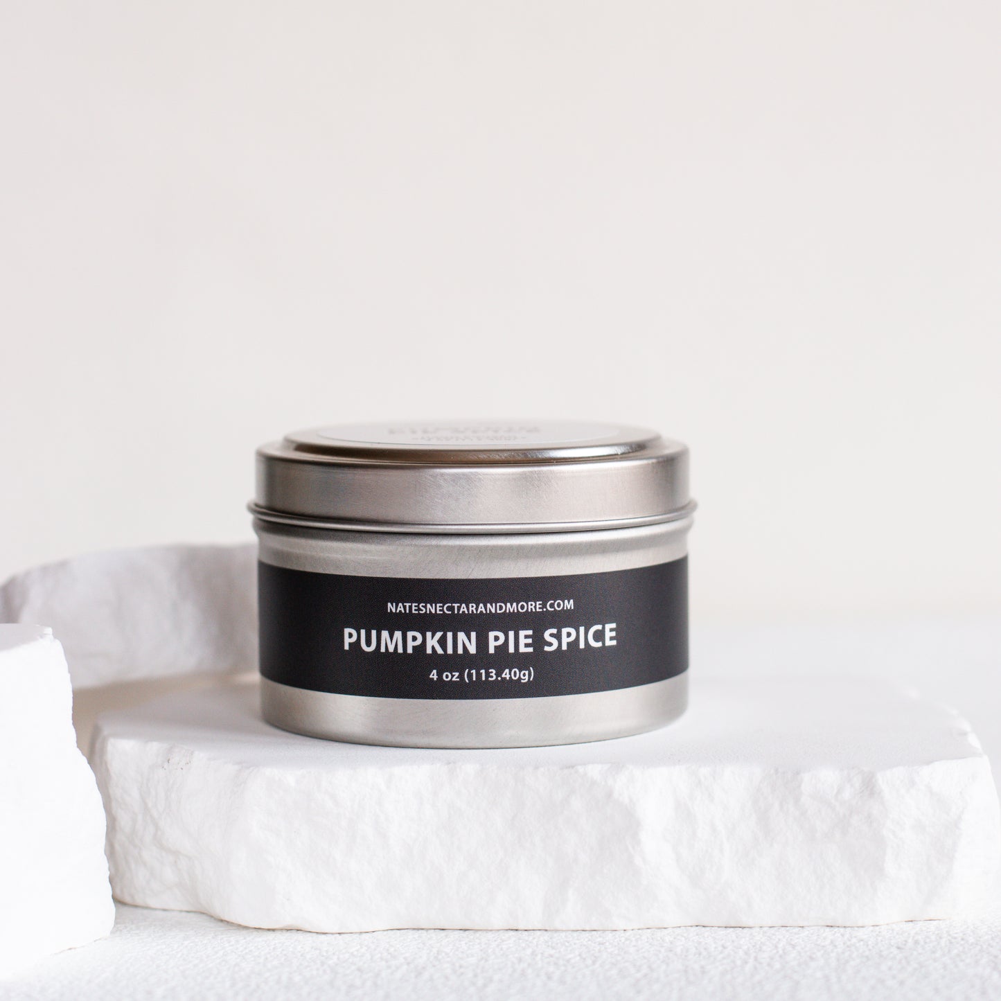 A silver metal tin candle labeled 'PUMPKIN PIE SPICE' by Natesh & Co, displayed on a white stone slab against a clean white background.