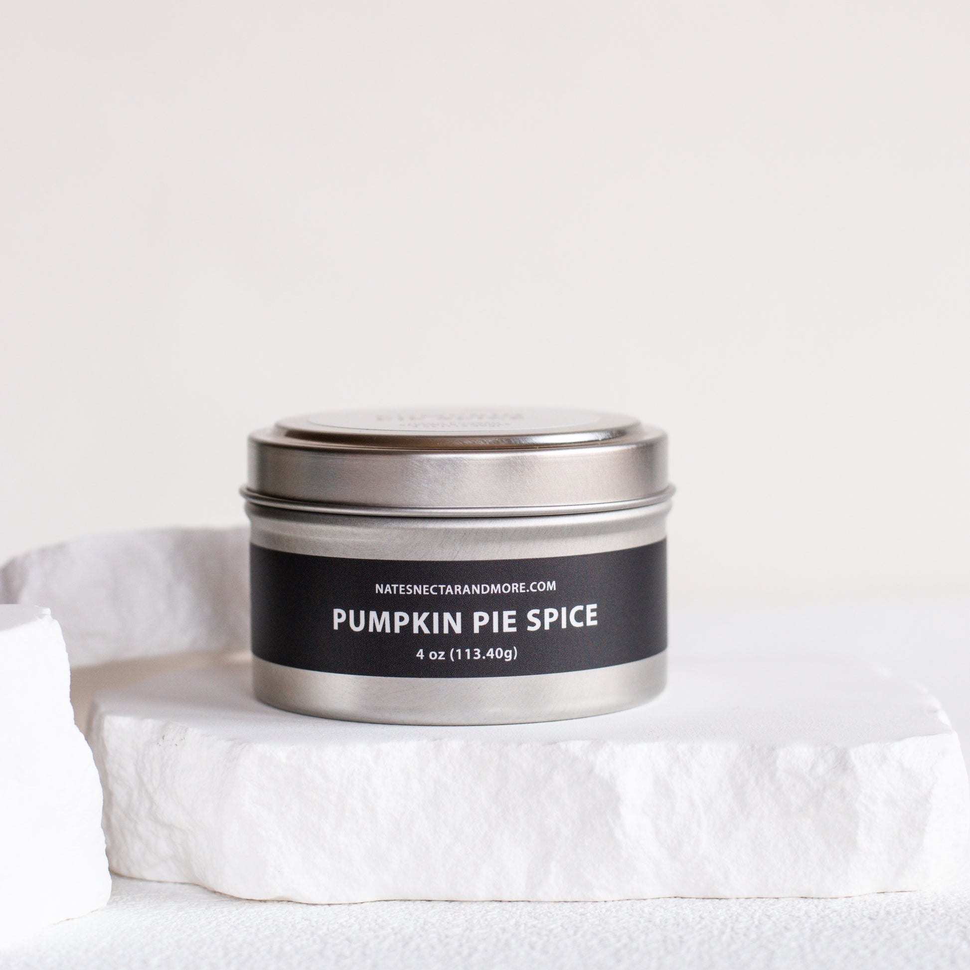 A silver metal tin candle labeled 'PUMPKIN PIE SPICE' by Natesh & Co, displayed on a white stone slab against a clean white background.