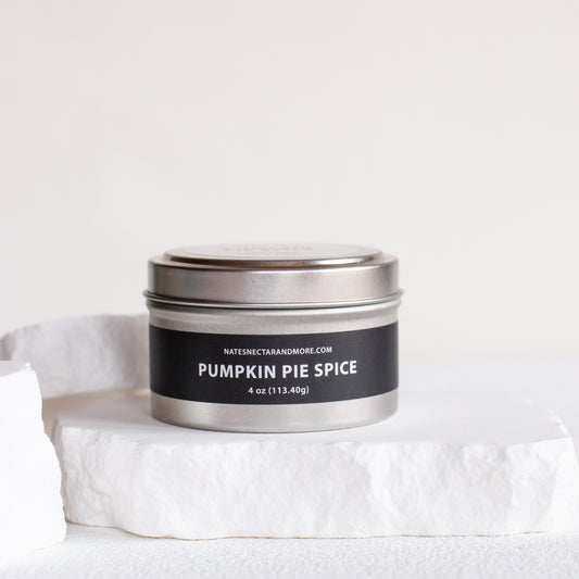 A silver metal tin candle labeled 'PUMPKIN PIE SPICE' by Natesh & Co, displayed on a white stone slab against a clean white background.