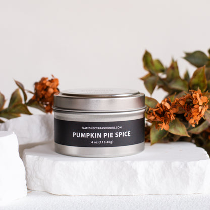 A silver metal tin candle labeled 'PUMPKIN PIE SPICE' by Nataniel Randomore, styled on a white stone slab with dried orange flowers in the background.
