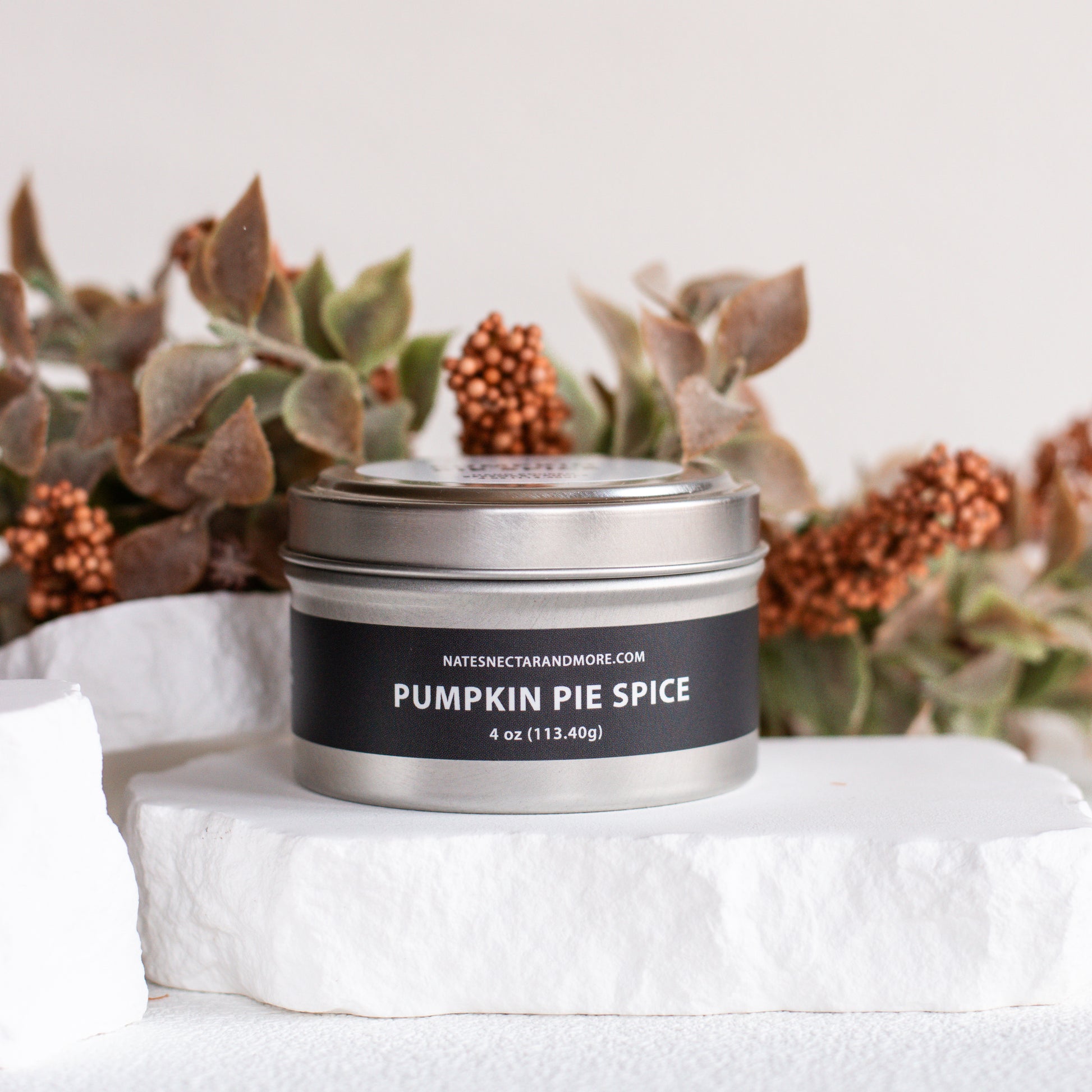A silver metal tin candle labeled 'PUMPKIN PIE SPICE' by Nate's Secret in a fall-themed setting with dried leaves and pinecones on a white stone pedestal.