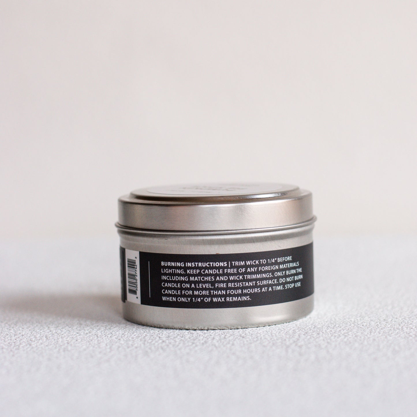 A small round metal tin candle shown from the back, with a black label containing white text and a barcode, set against a plain light background.