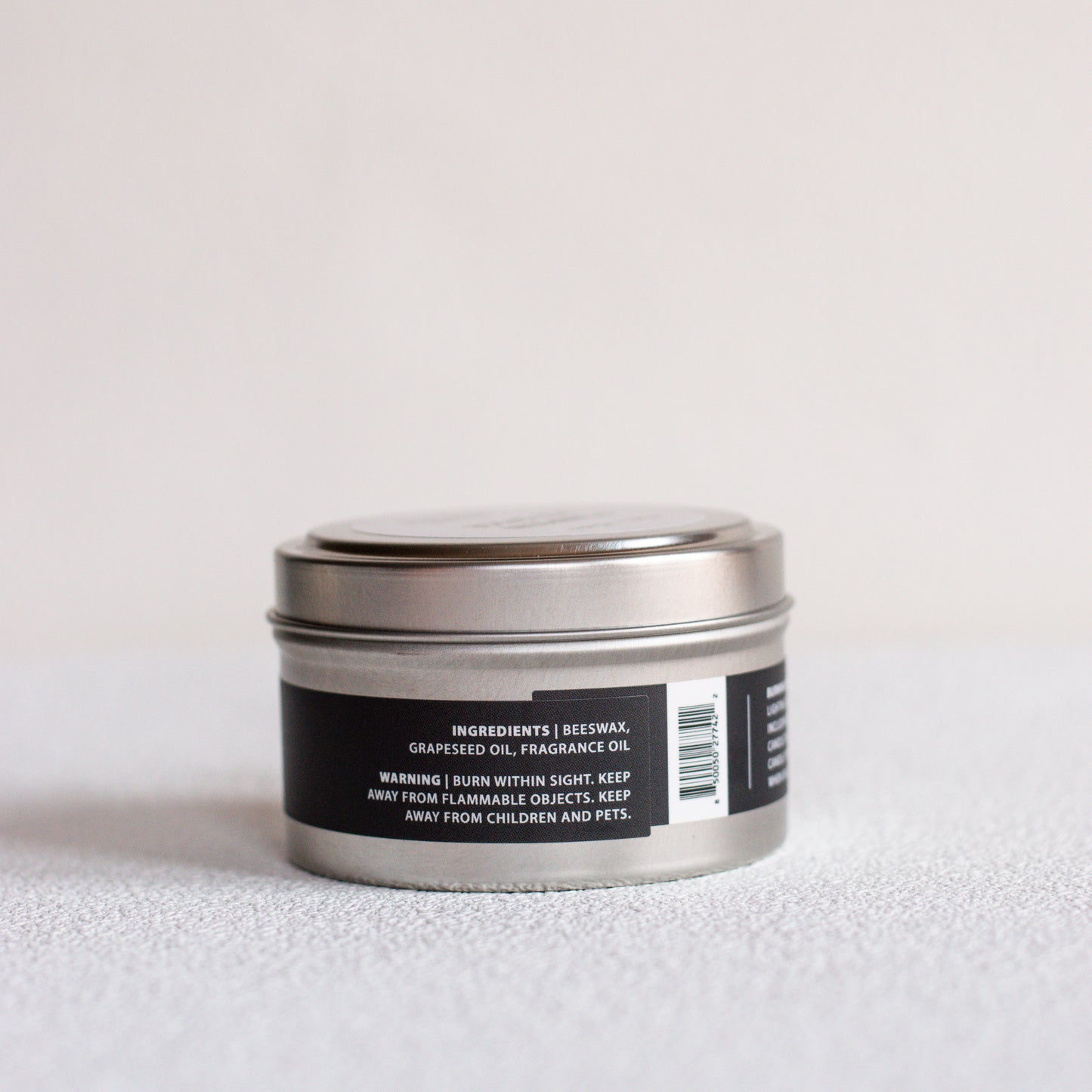 A small round metal tin candle with a silver lid and a black label, photographed on a light neutral surface against a pale background.